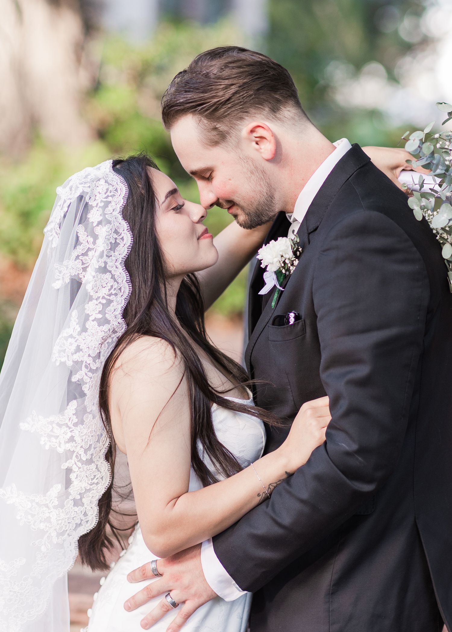 Bride in a lace-trimmed veil and groom in a black suit with a white boutonniere share a tender embrace outdoors on their wedding day.