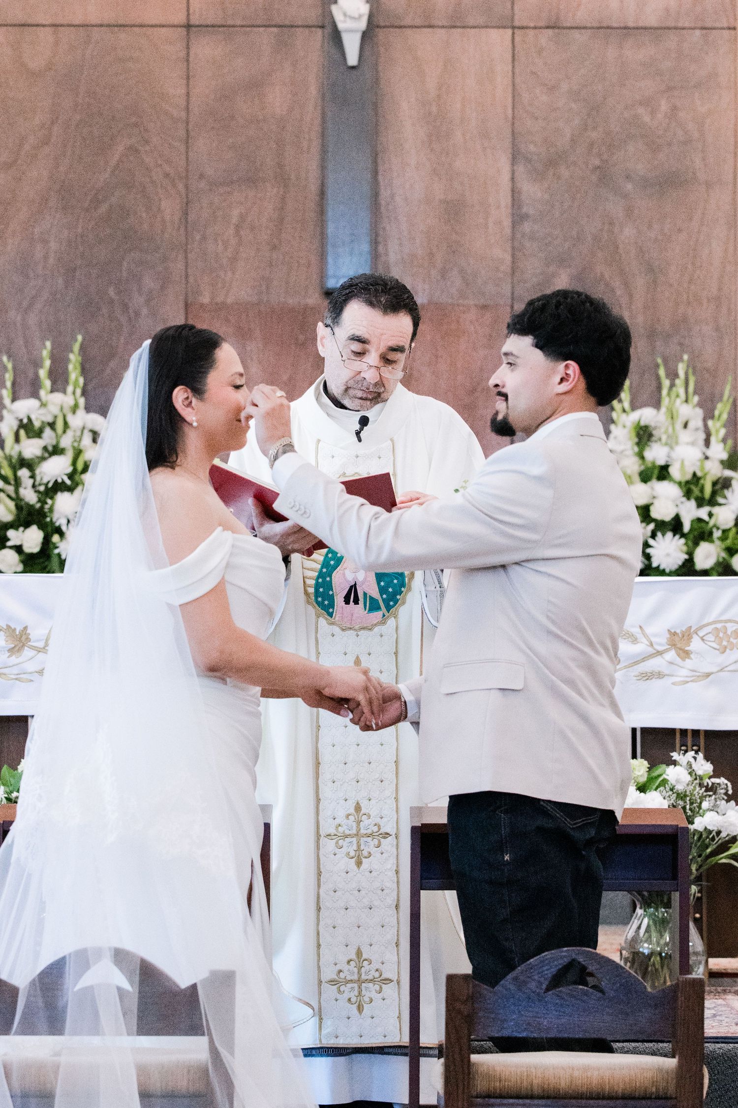 Bride and groom laugh joyfully while exchanging vows before smiling priest at church altar.