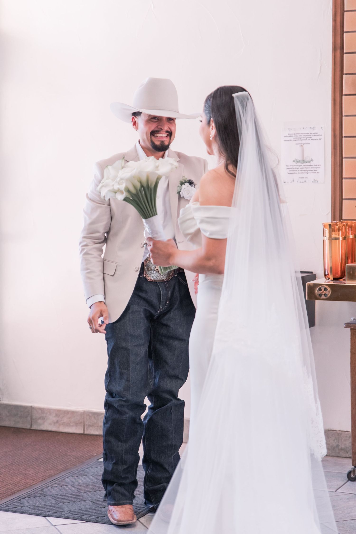 Groom in cowboy hat and beige suit smiles at bride in white off-shoulder gown at church entrance.