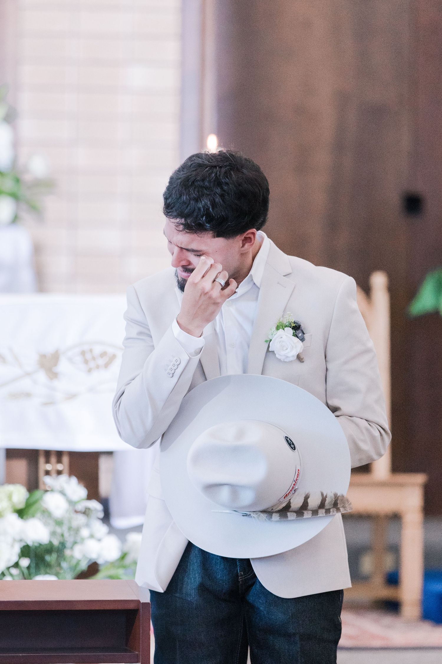 Tearful groom in beige suit holds cowboy hat while wiping eyes at church altar during wedding.