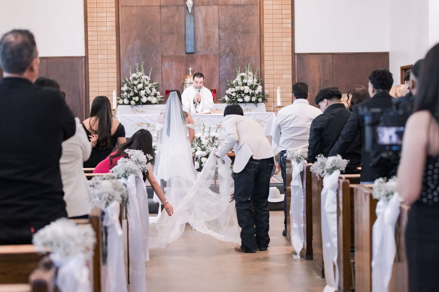 Wedding party arranges bride's veil train in decorated Catholic church before ceremony begins.