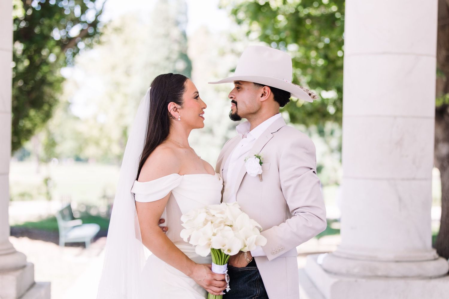 Bride holding white rose bouquet gazes at groom in tan suit and cowboy hat between white columns.