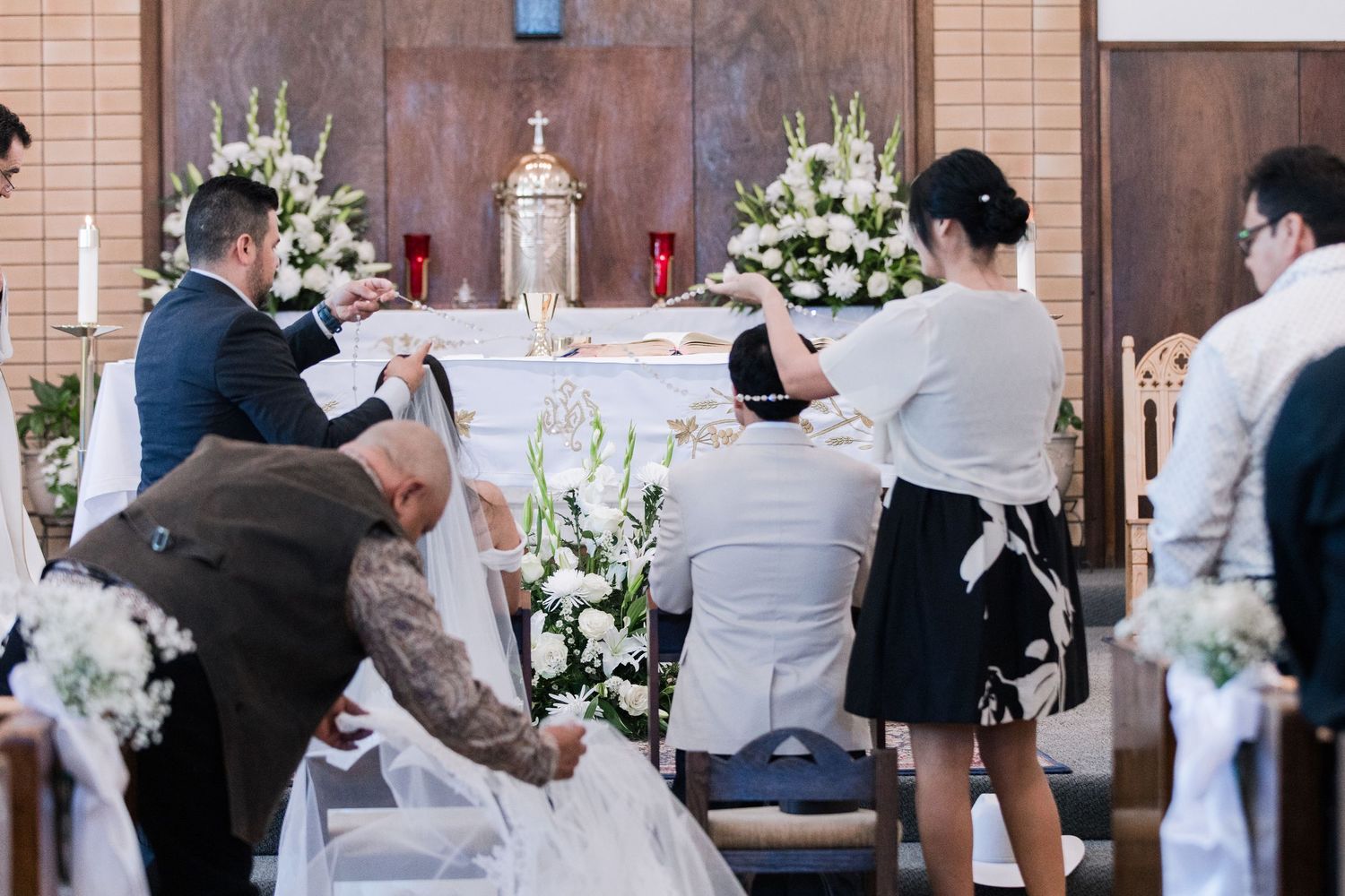 Guests and wedding party gather around the altar helping the bride during a Catholic wedding ceremony.