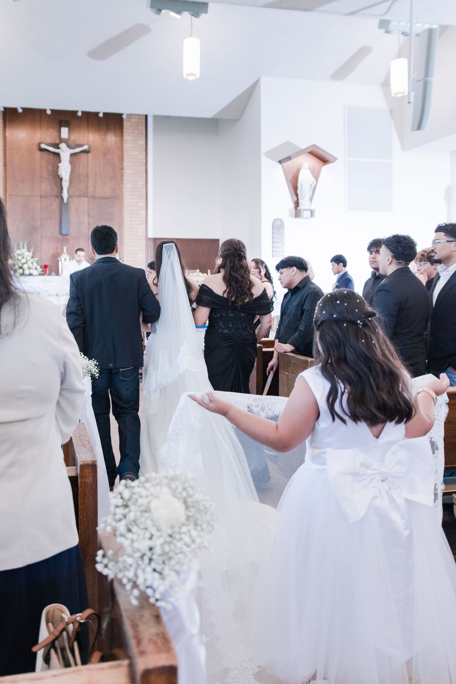 Flower girl in white dress holds bouquet as bride and groom stand at altar in Catholic church.