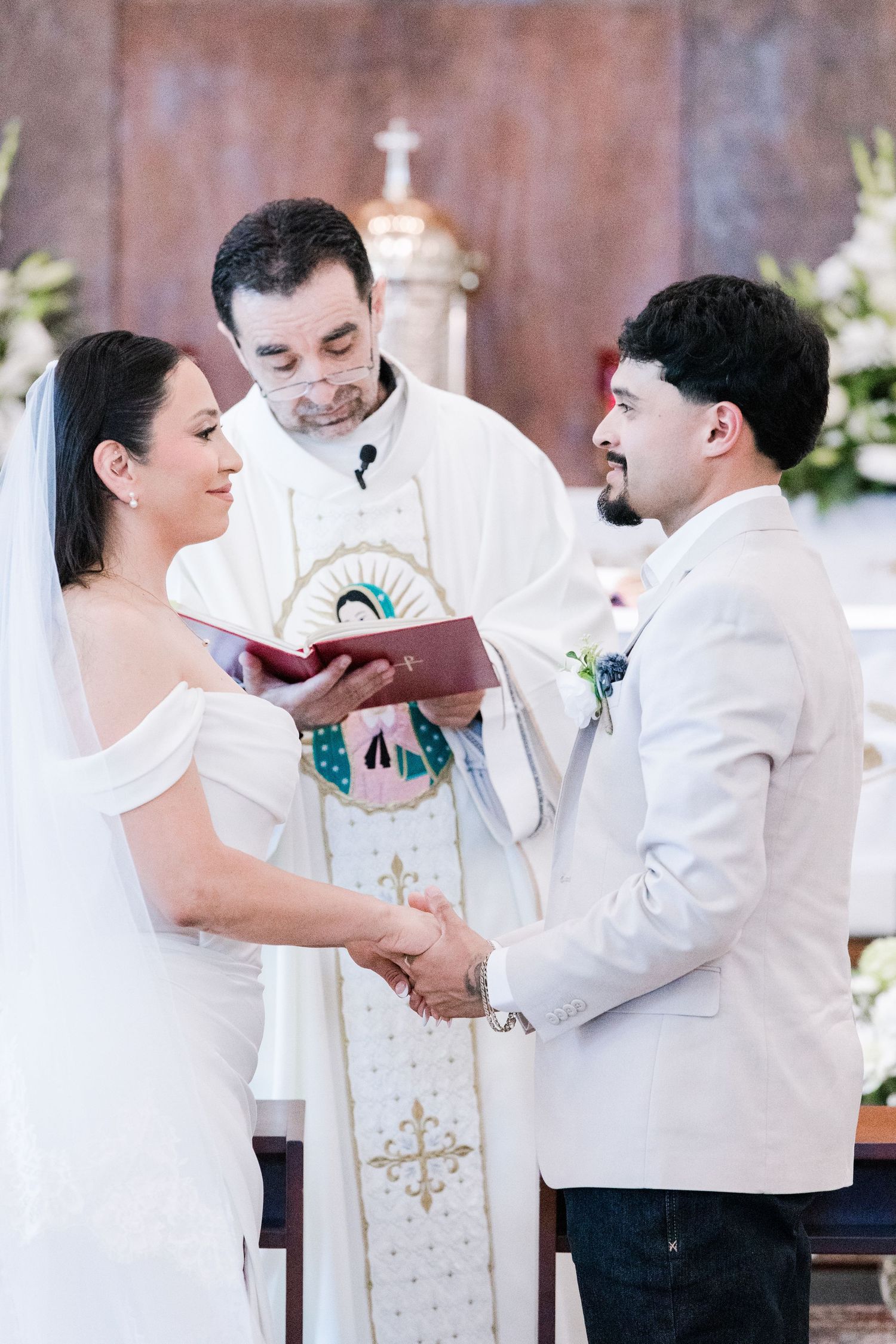 Bride and groom hold hands exchanging vows before priest at floral-decorated Catholic church altar.