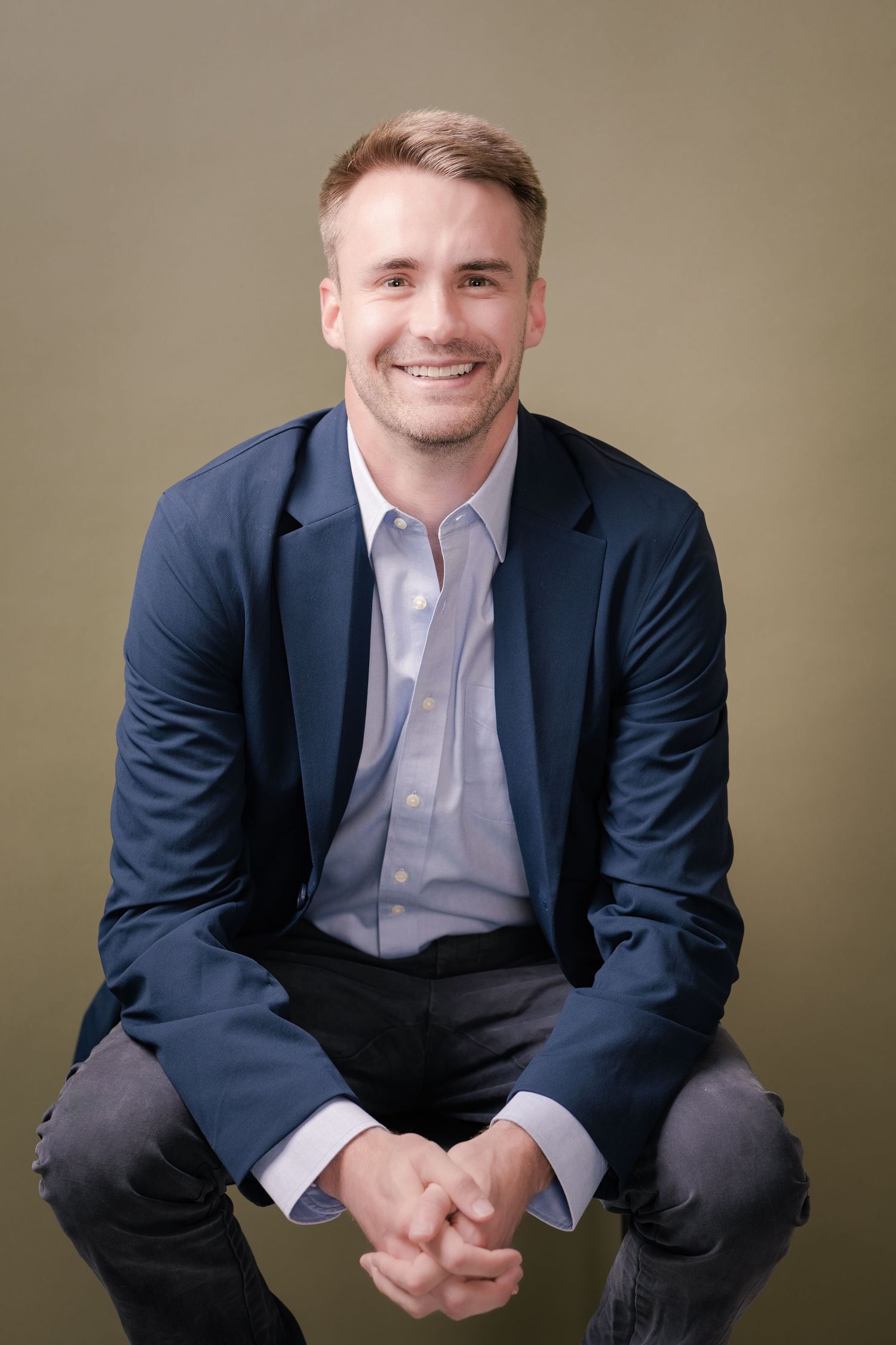 Smiling man with short hair in a navy blazer and light blue shirt sits with clasped hands against an olive green studio backdrop.