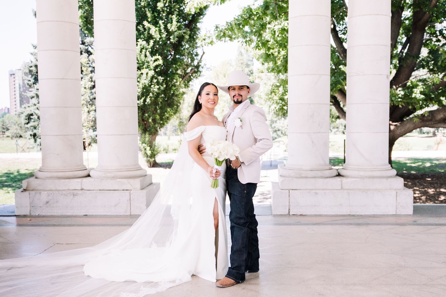 Bride and groom smile together under white stone columns, her long cathedral veil flowing behind her.
