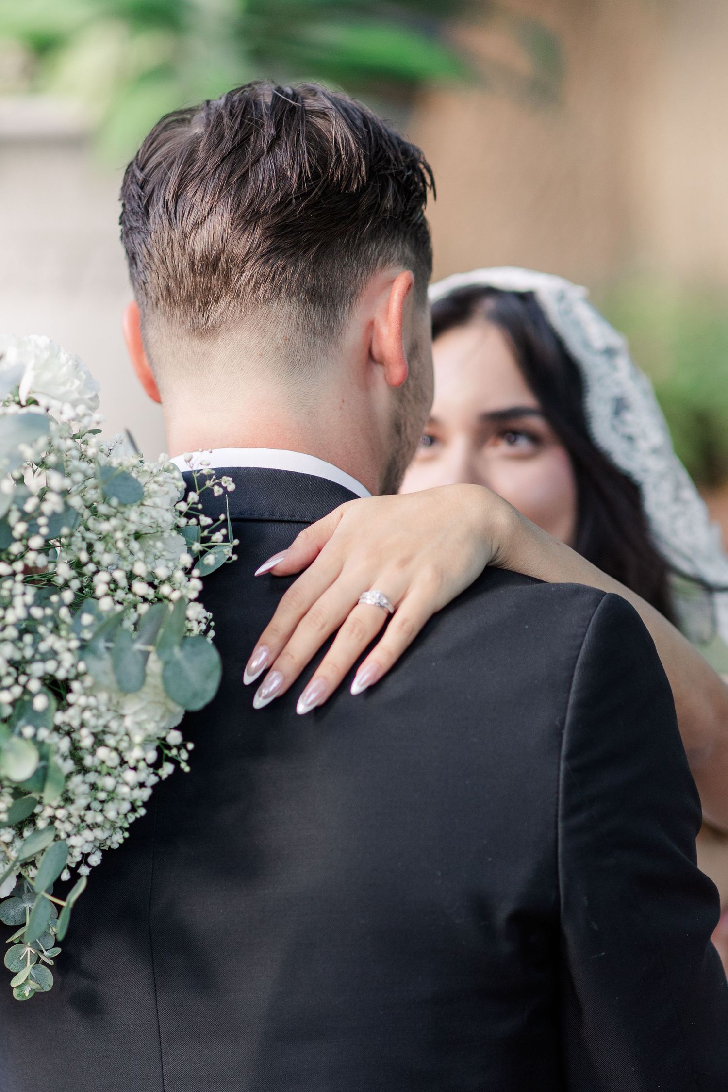 Bride in a lace veil embraces groom in a black suit, her manicured hand and diamond ring on his shoulder beside a eucalyptus bouquet.