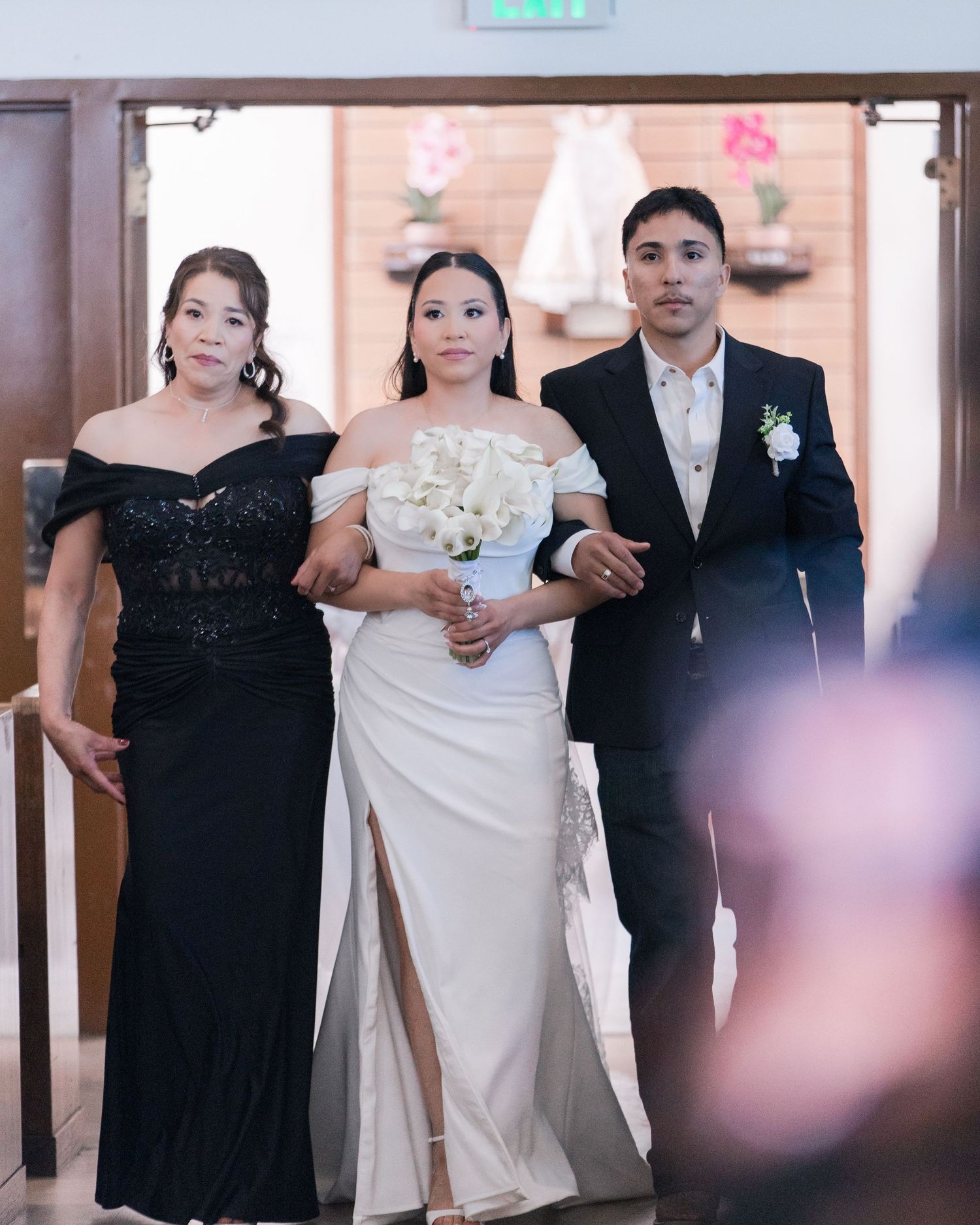 Bride in white off-shoulder gown walks down aisle escorted by parents at church wedding ceremony.