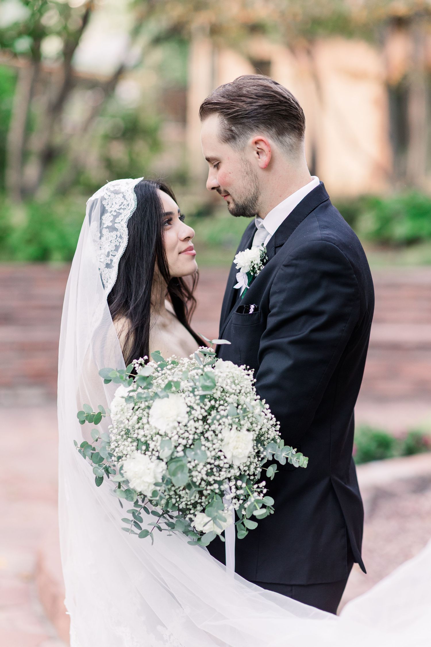 Bride in a lace-trimmed veil holding a white baby's breath bouquet gazes at a groom in a dark suit in a garden.