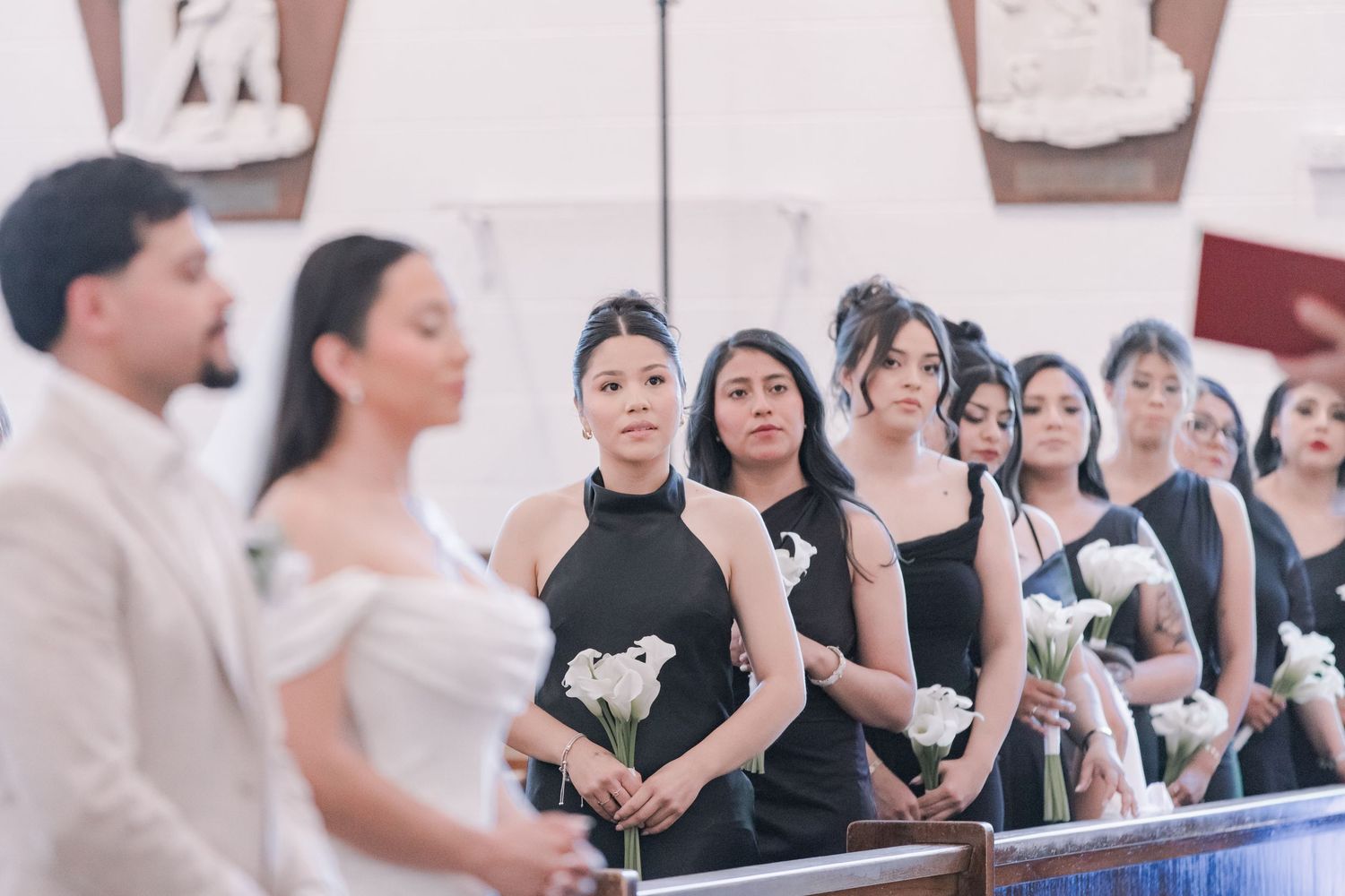 Bridesmaids in black dresses holding white calla lilies stand attentively during a church wedding ceremony.