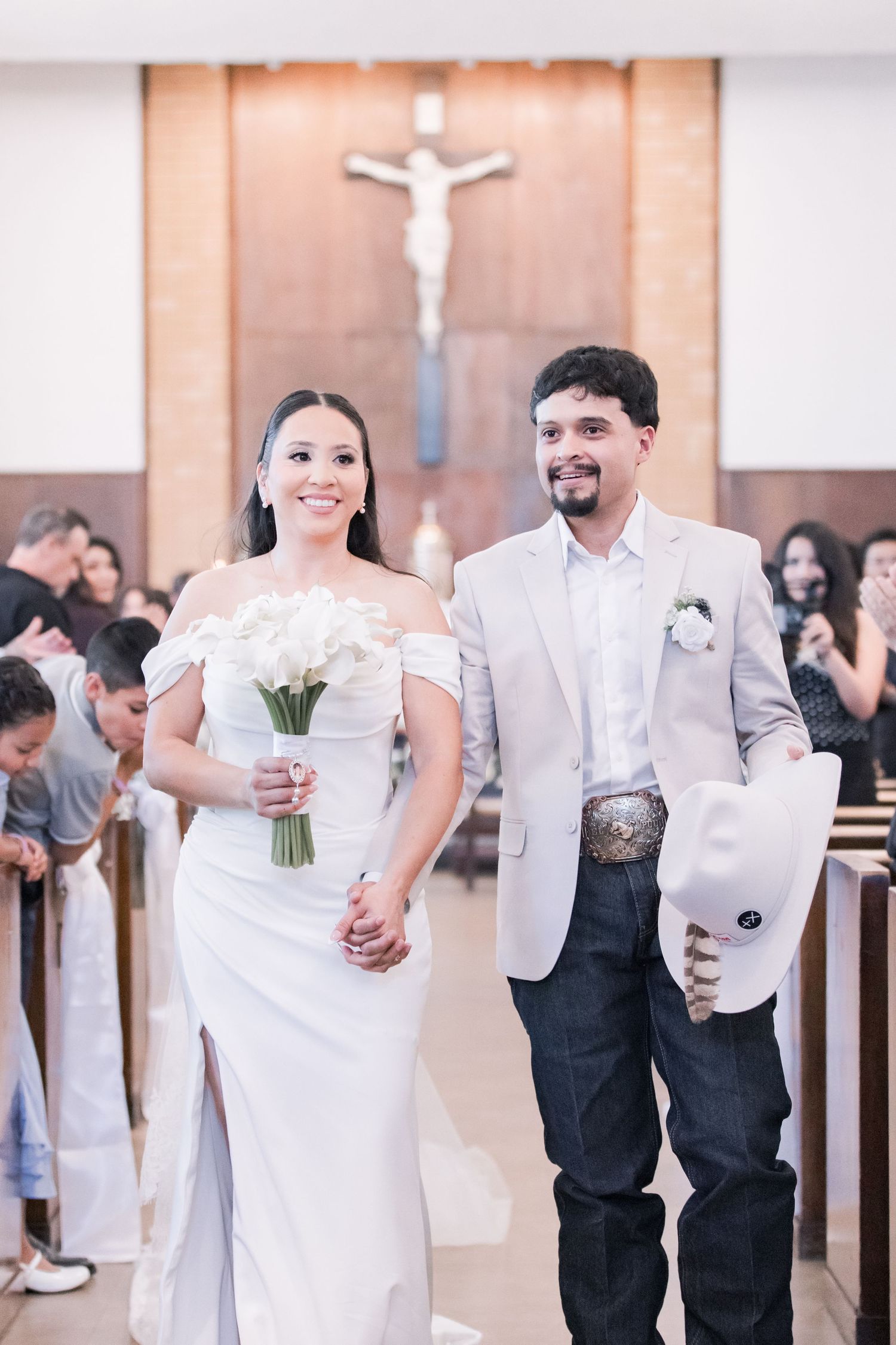 Happy newlyweds walk down church aisle, groom in beige suit holding cowboy hat, bride with white bouquet.
