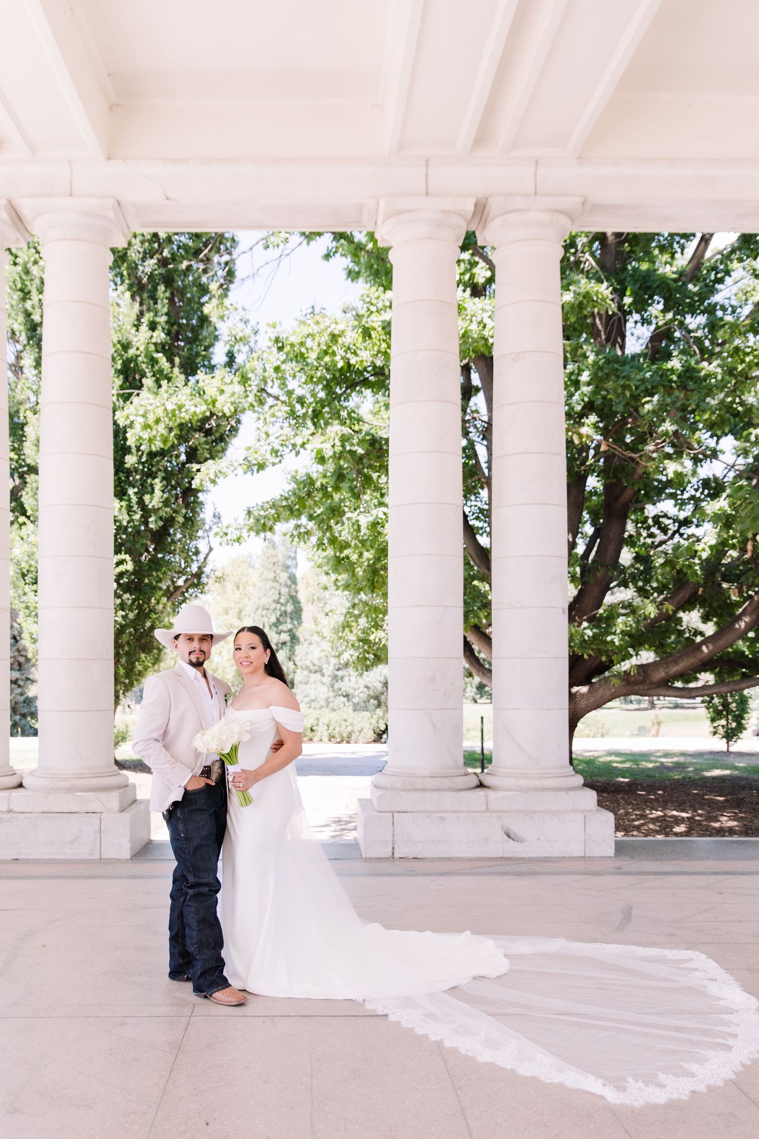 Bride and groom pose under white columns in a park, her cathedral veil trailing elegantly on the ground.