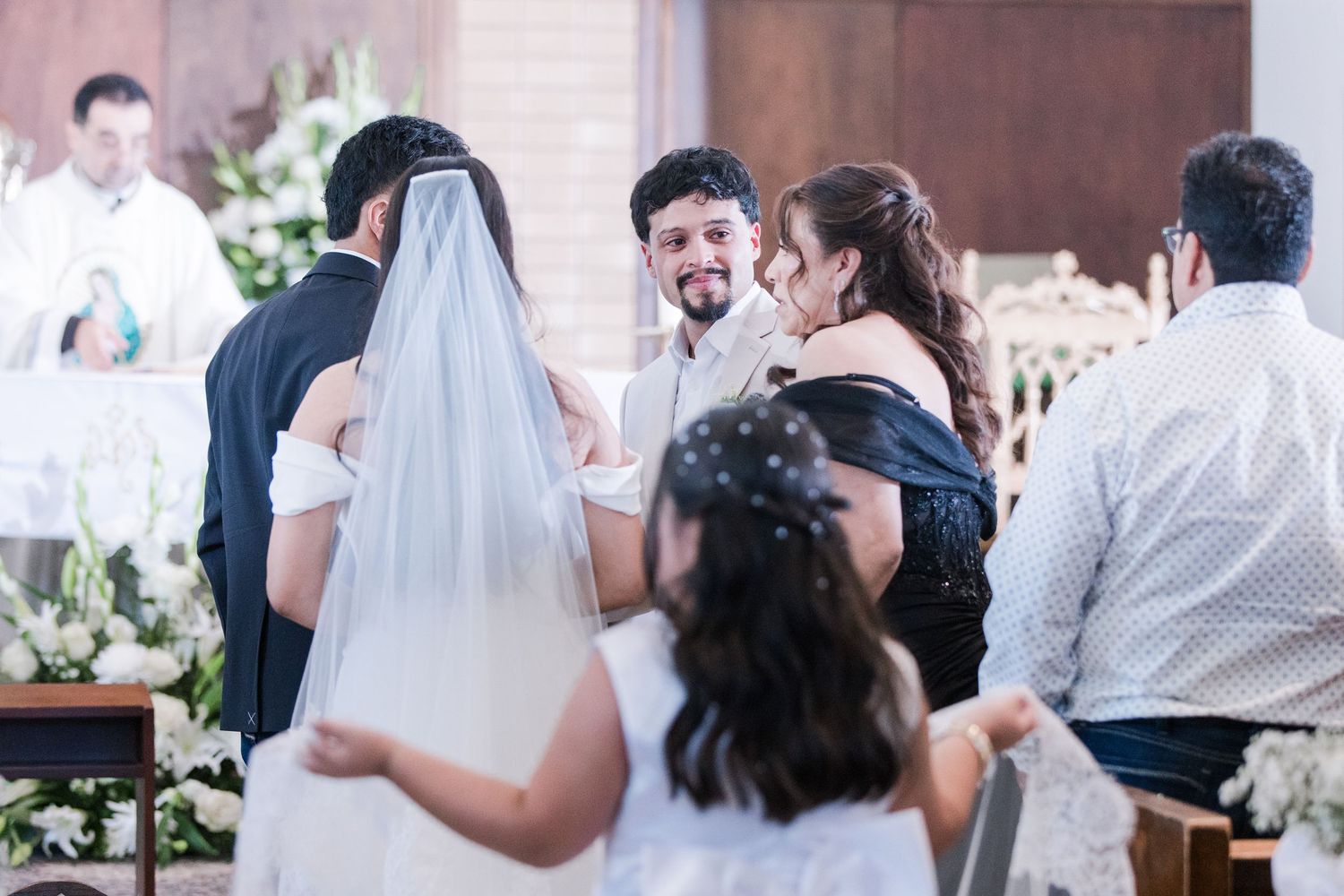 Smiling groom in white suit greets guests at church altar during wedding ceremony.
