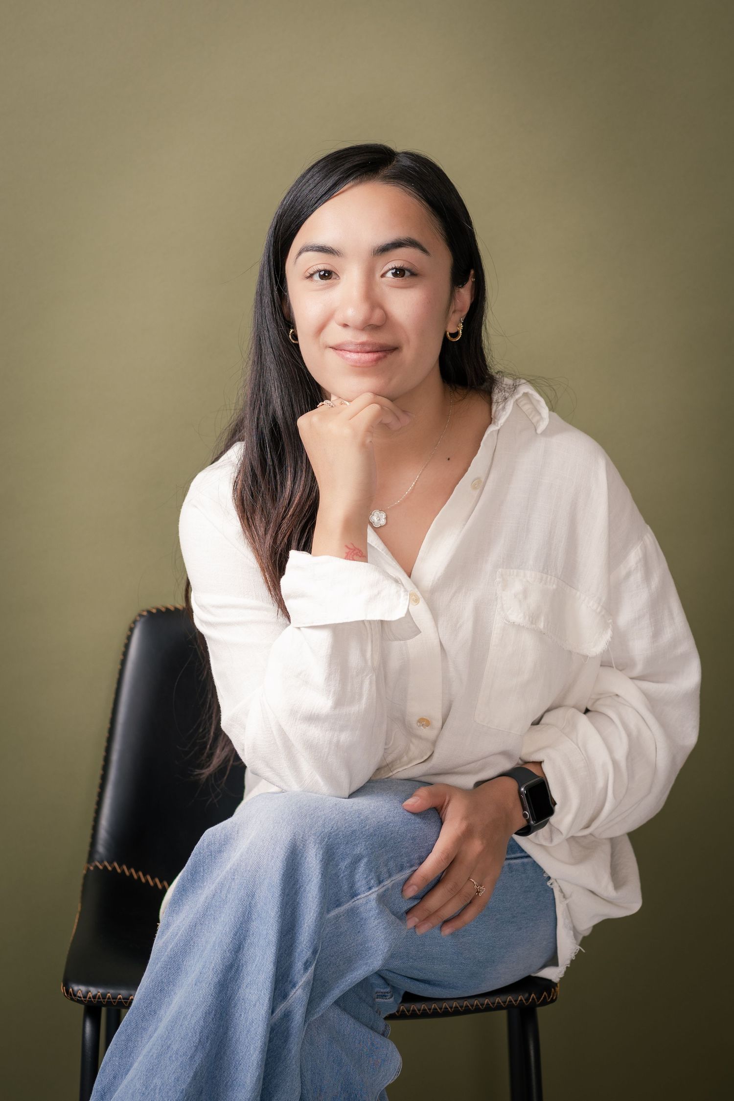 Smiling woman with long dark hair sits on a black chair, resting her chin on her hand, in a white shirt and jeans against an olive wall.