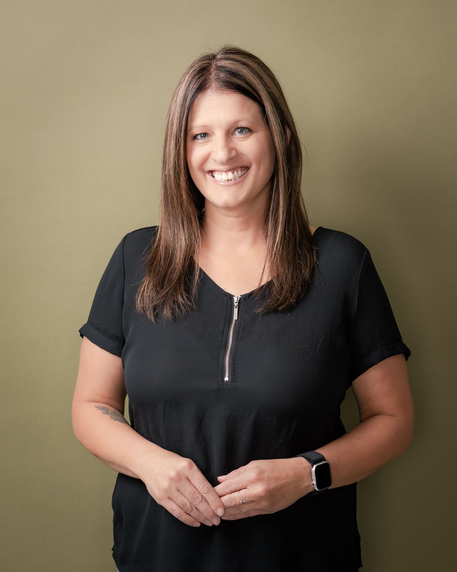 Smiling woman with long brown highlighted hair wearing a black zip-neck top and smartwatch, hands clasped against an olive green background.