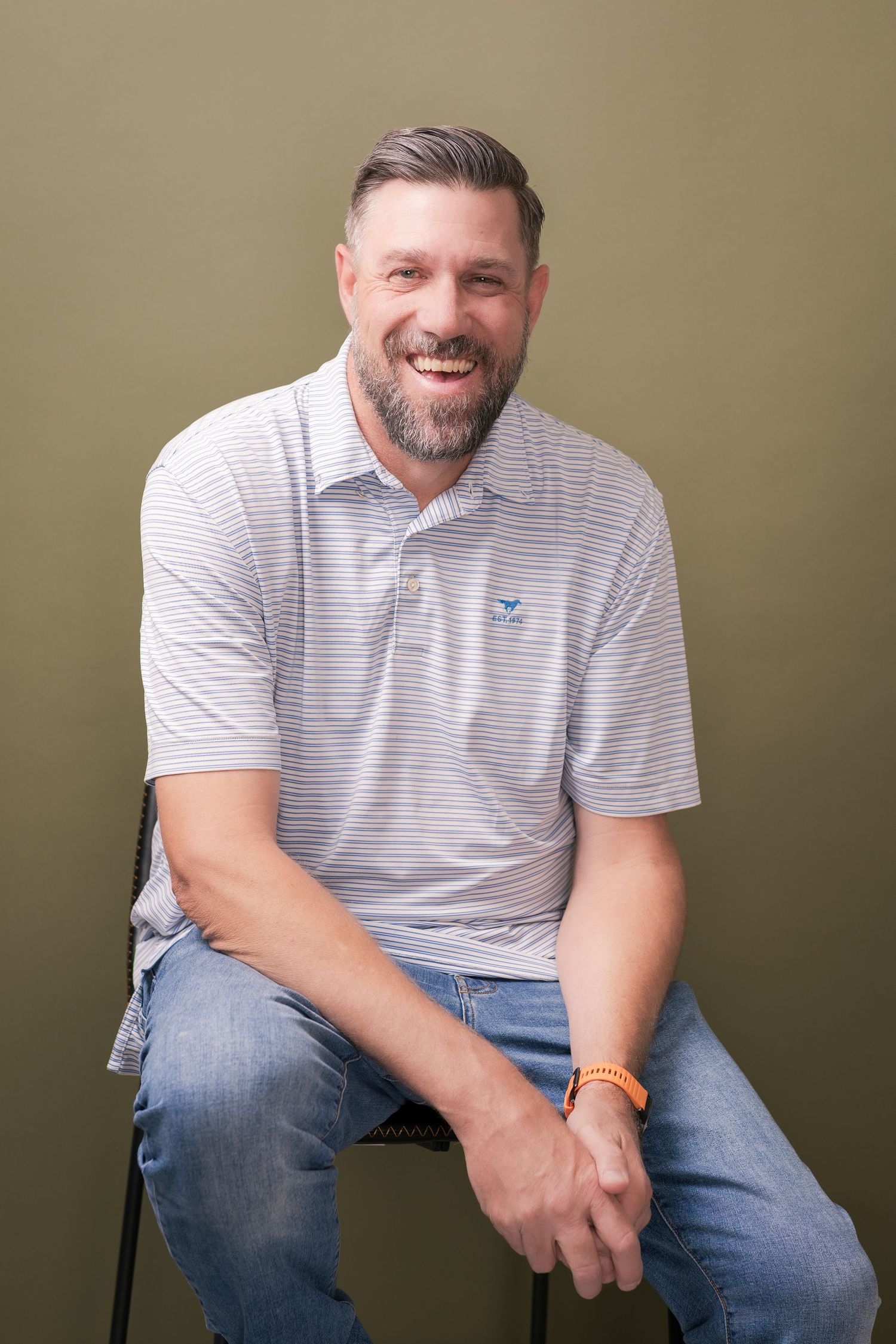 Smiling bearded man in a striped polo shirt and jeans sits on a stool against an olive green backdrop, wearing an orange wristband.
