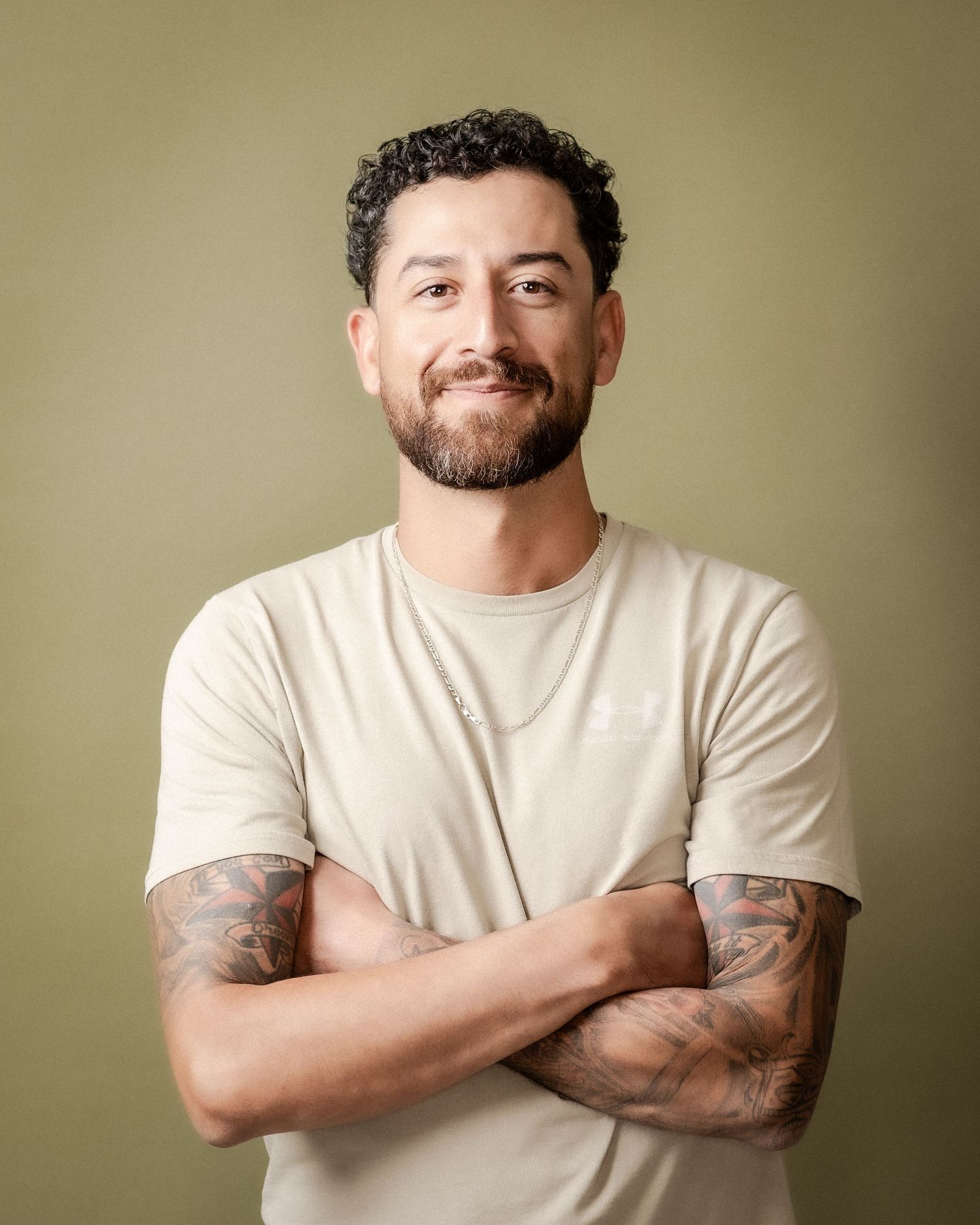 Smiling bearded man with curly dark hair and tattooed forearms stands with arms crossed in a beige t-shirt against an olive green backdrop.