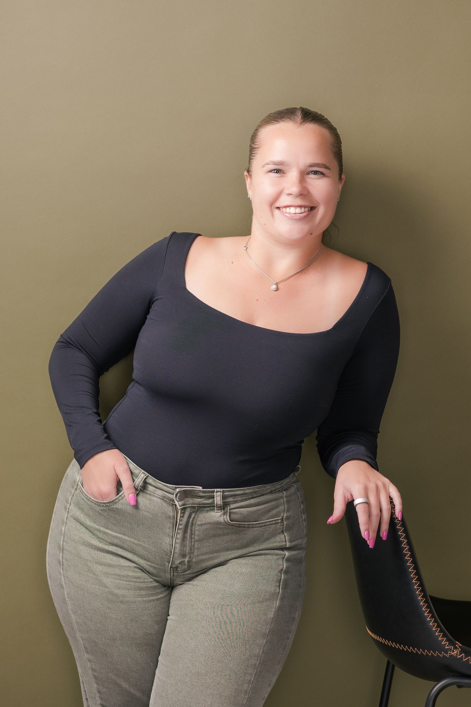 Smiling woman in a black scoop-neck top and olive jeans leans casually on a black leather bar stool against an olive green wall.