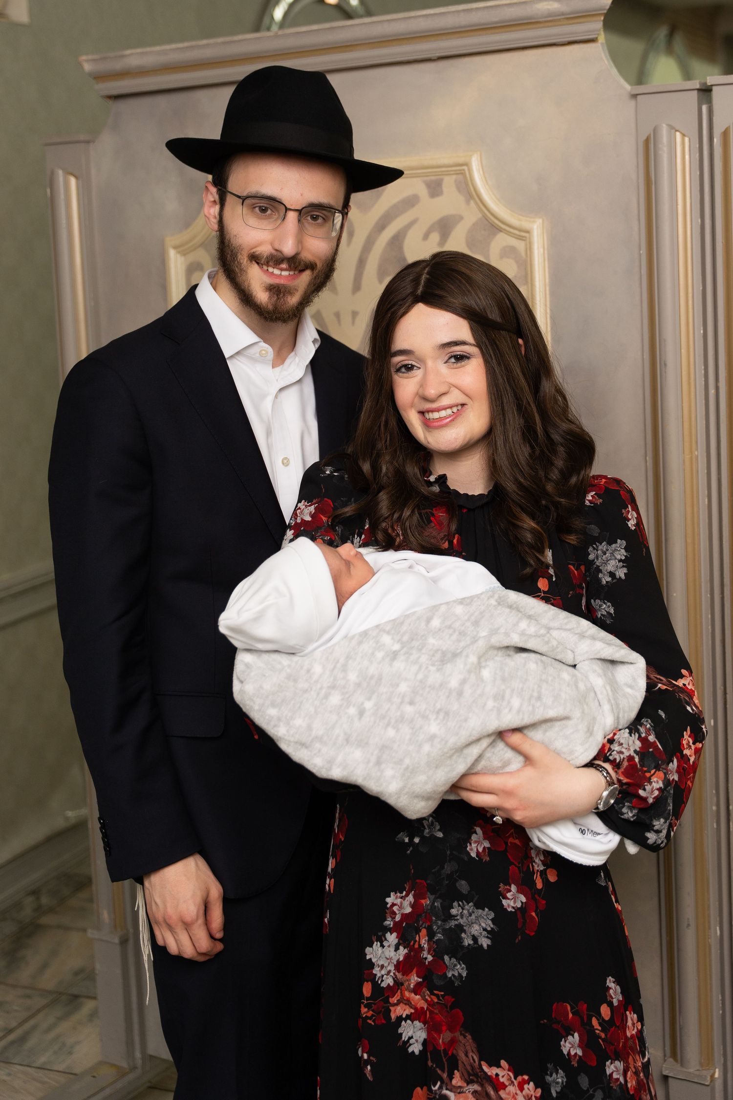 A couple in formal attire stands together while holding a baby wrapped in white blankets.