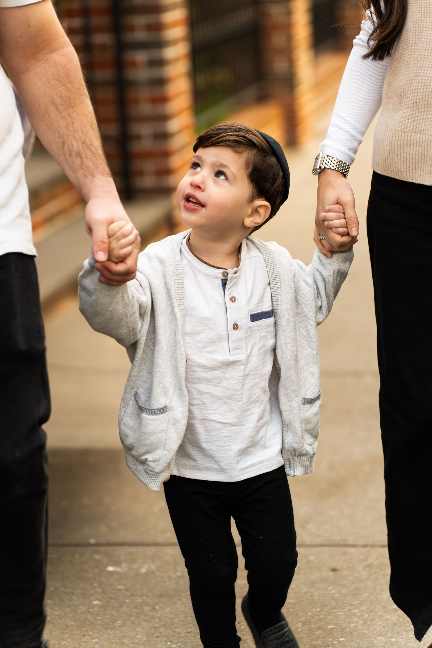 Small child walking while holding hands with adults on brick-lined sidewalk.