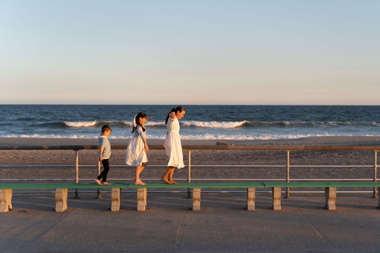 Three people in white dresses walk along a beachfront railing at sunset with ocean waves in the background.