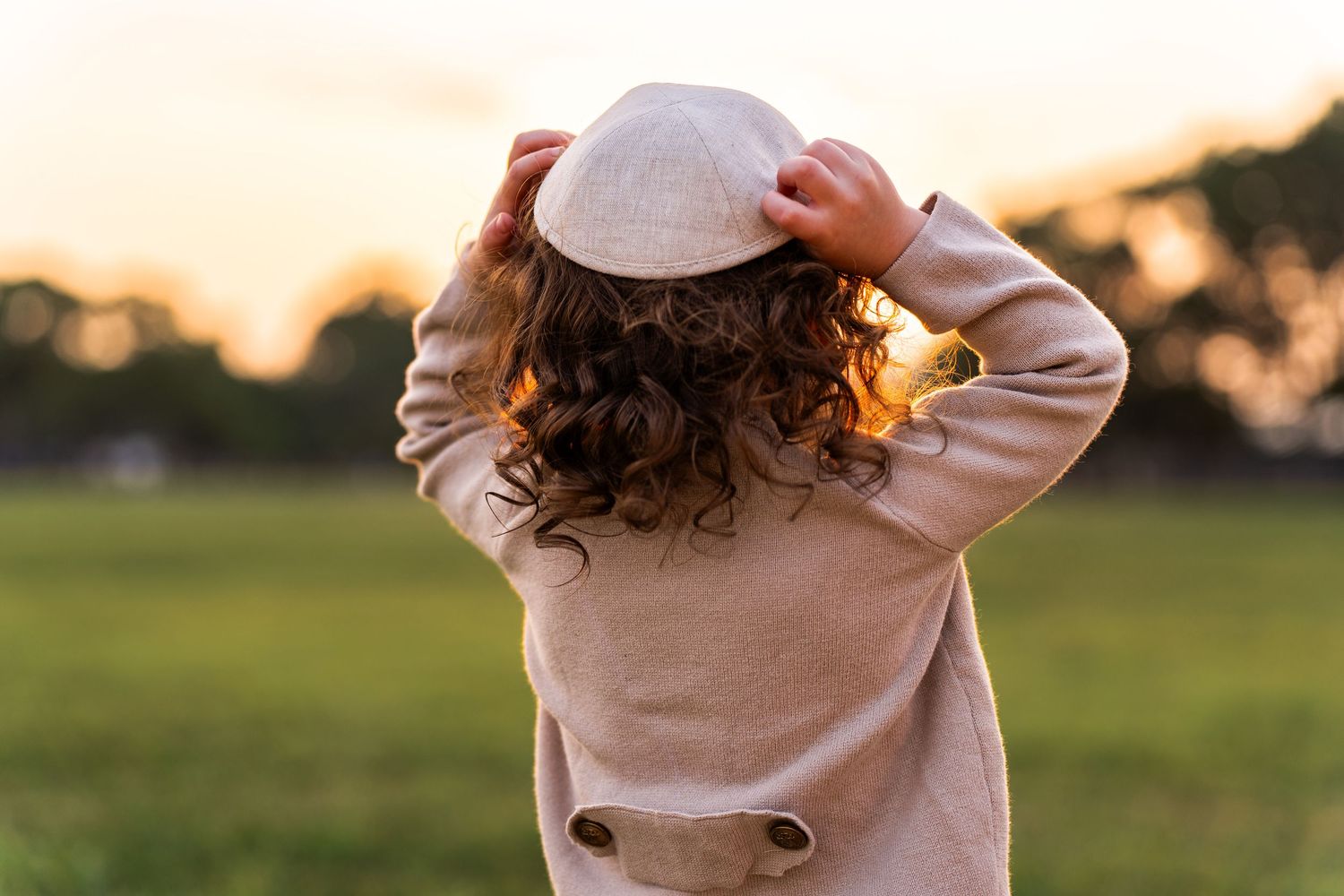 Person wearing a beige sweater stands in a grassy field at sunset with warm golden lighting.