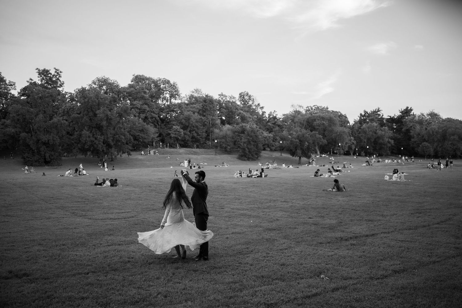 Couple dancing in a vast green park field with trees and people relaxing in black and white photo.