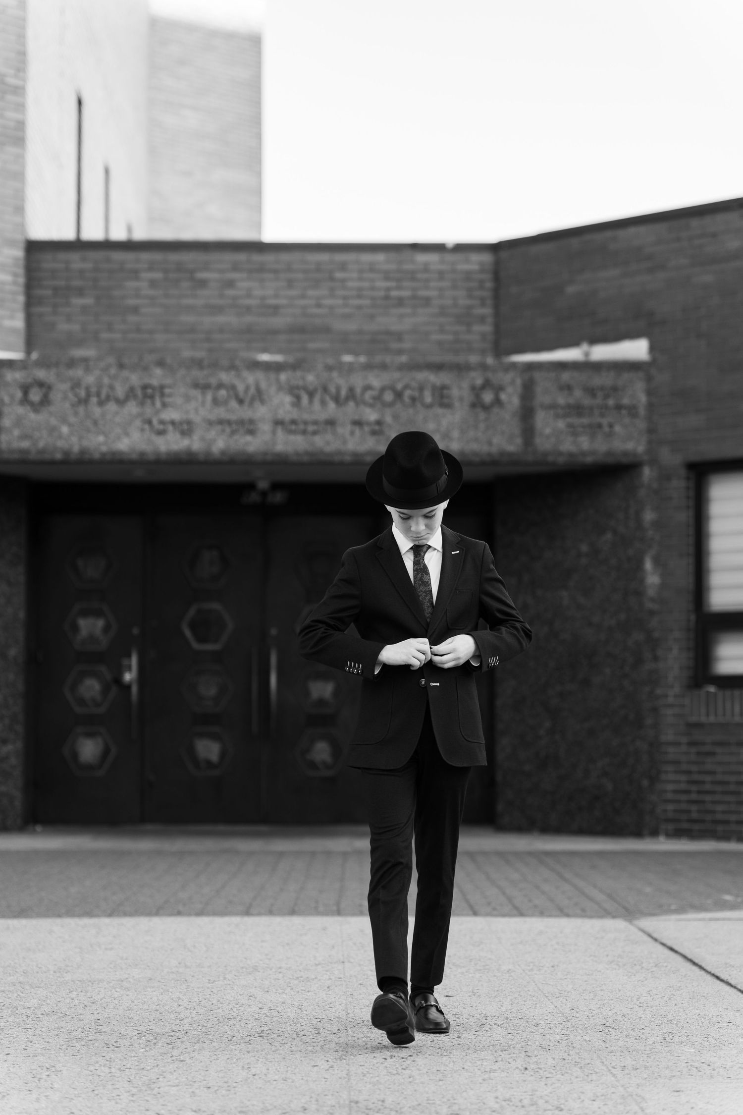 Person in formal attire stands alone outside brick synagogue building in black and white photo.