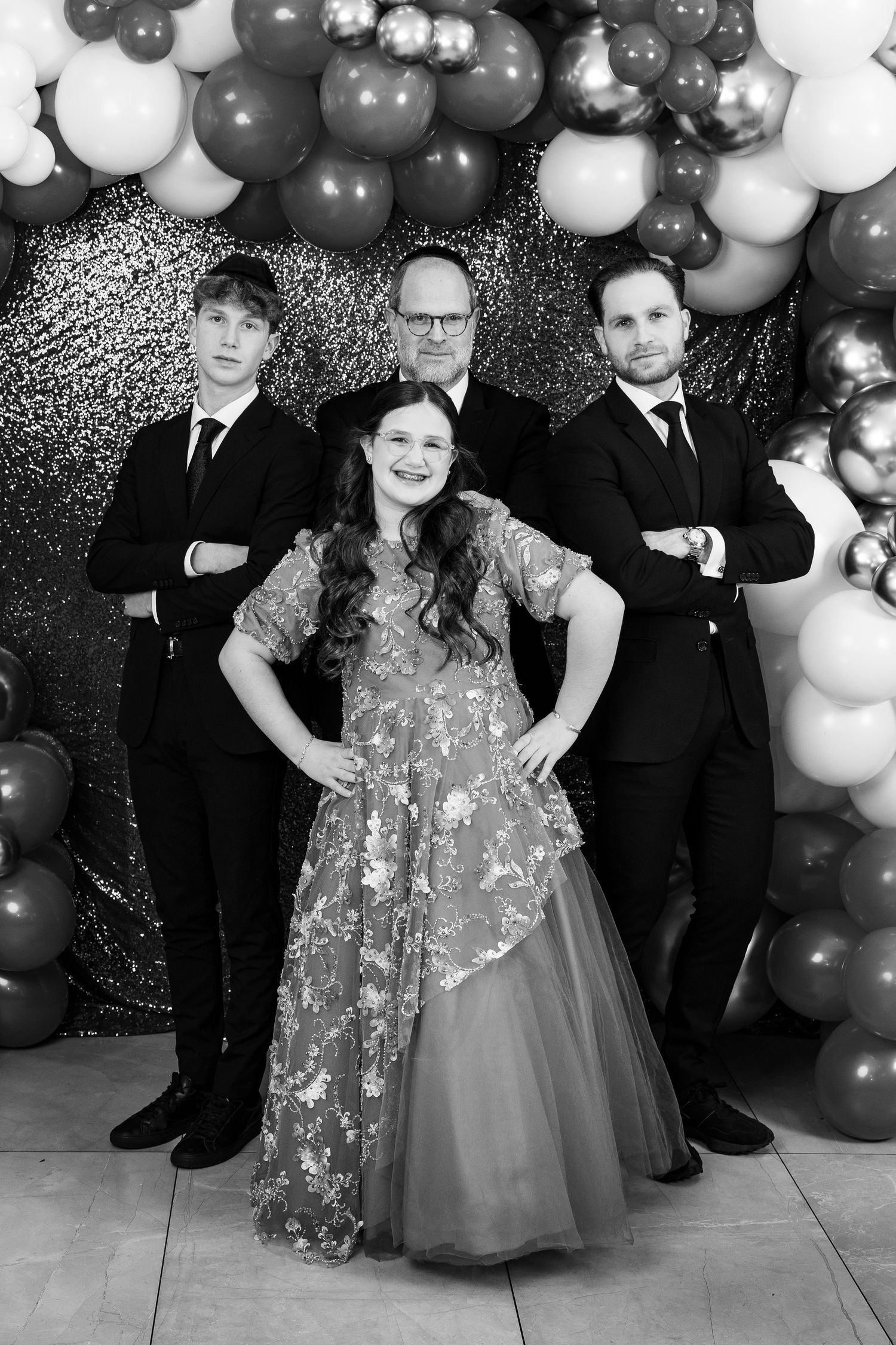 Black and white family portrait with woman in gown and three men in suits posed before balloon backdrop.
