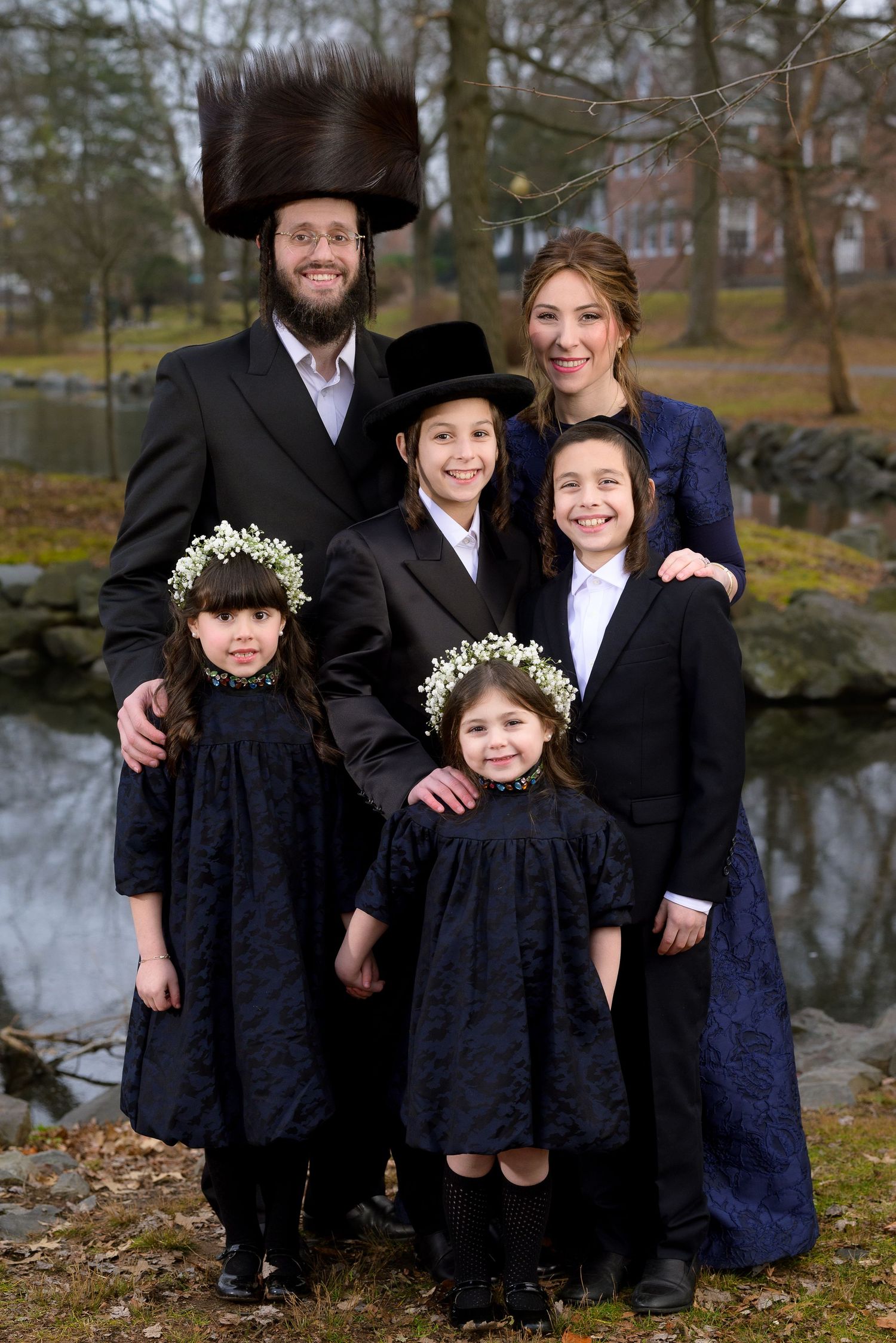 A formal family portrait by a pond featuring traditional Orthodox Jewish attire and floral headpieces.