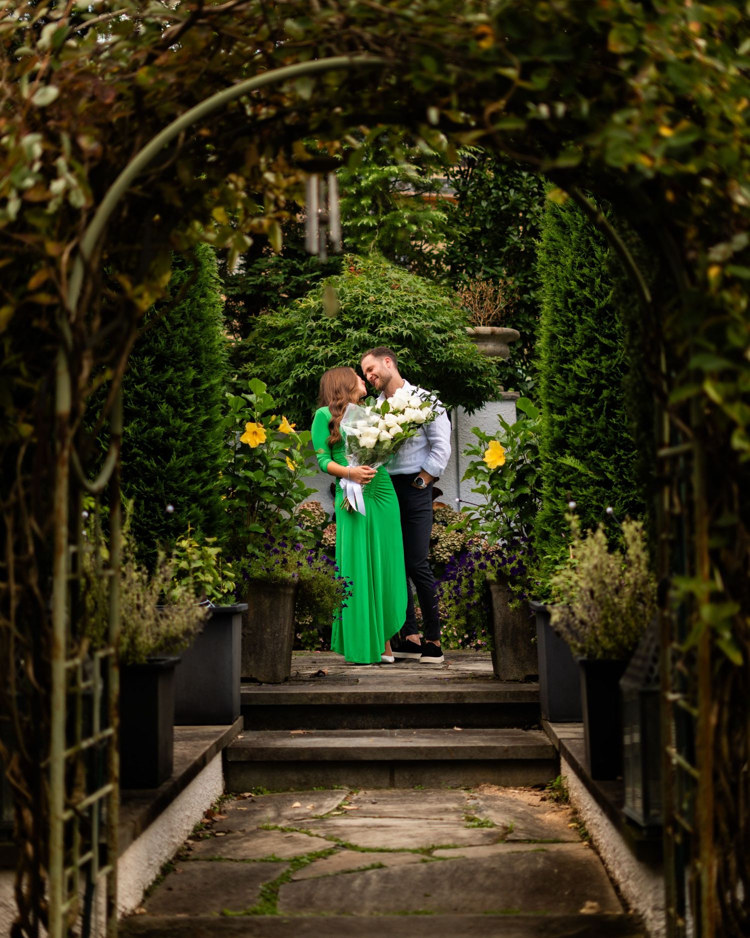 A couple embraces under a lush garden archway, with the person wearing an emerald green dress holding white flowers.