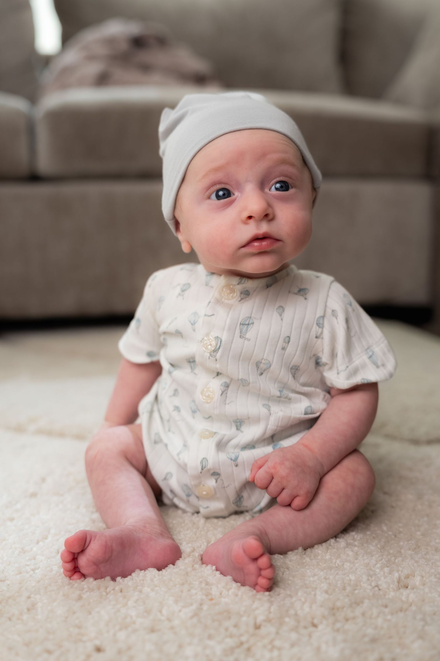 Adorable baby in gray hat and floral romper sitting on white carpet with couch in background.