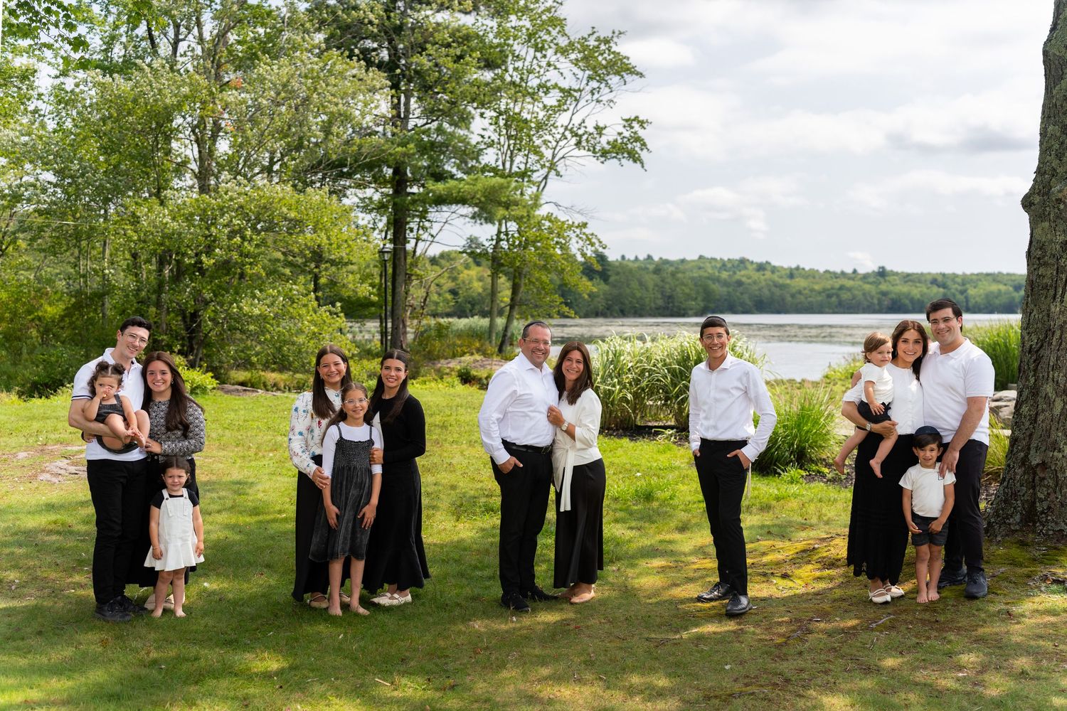 Large family portrait outdoors near a lake, dressed in black and white formal attire on a green lawn.