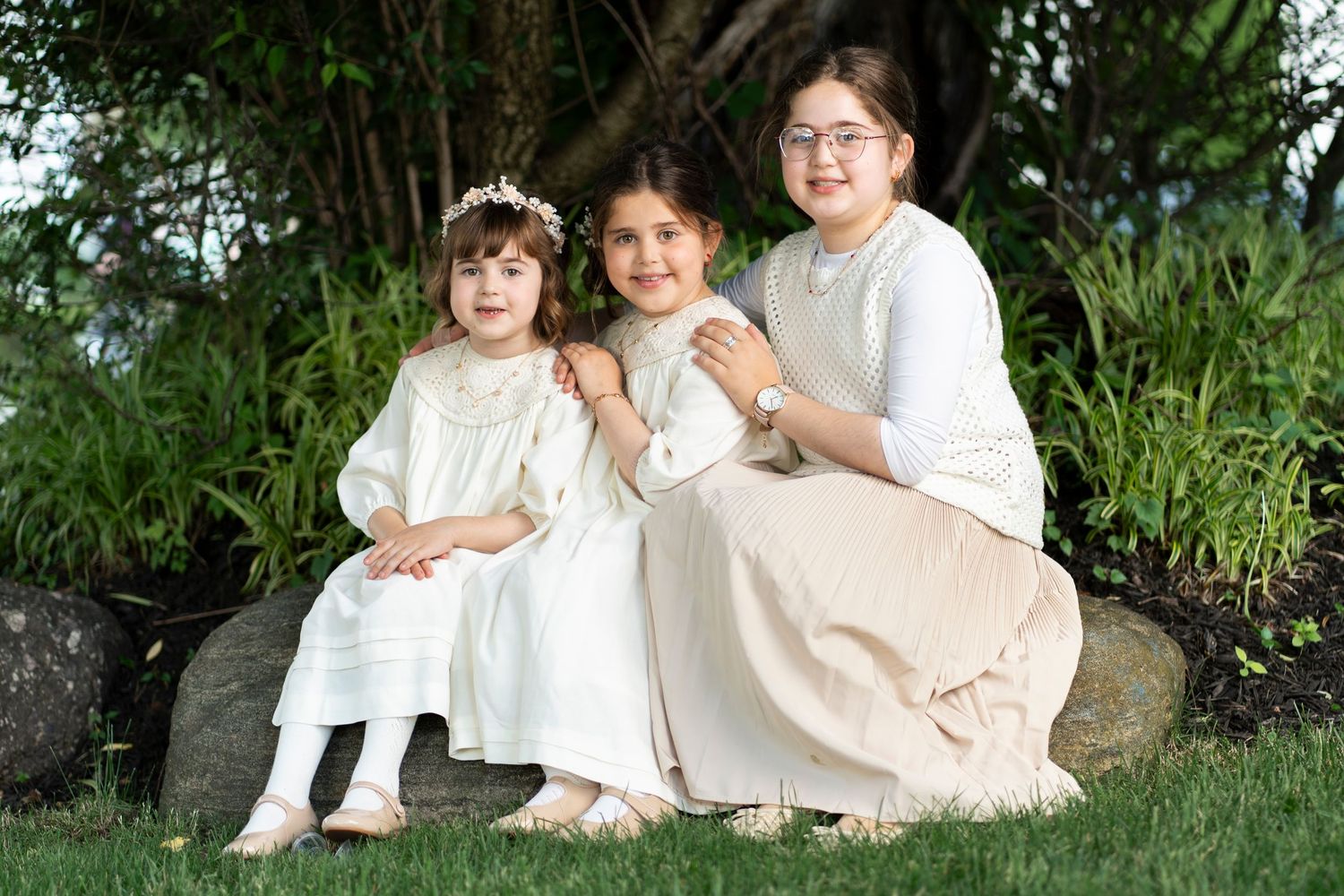 Children sitting together on grassy hill wearing coordinated white outfits in outdoor garden setting.