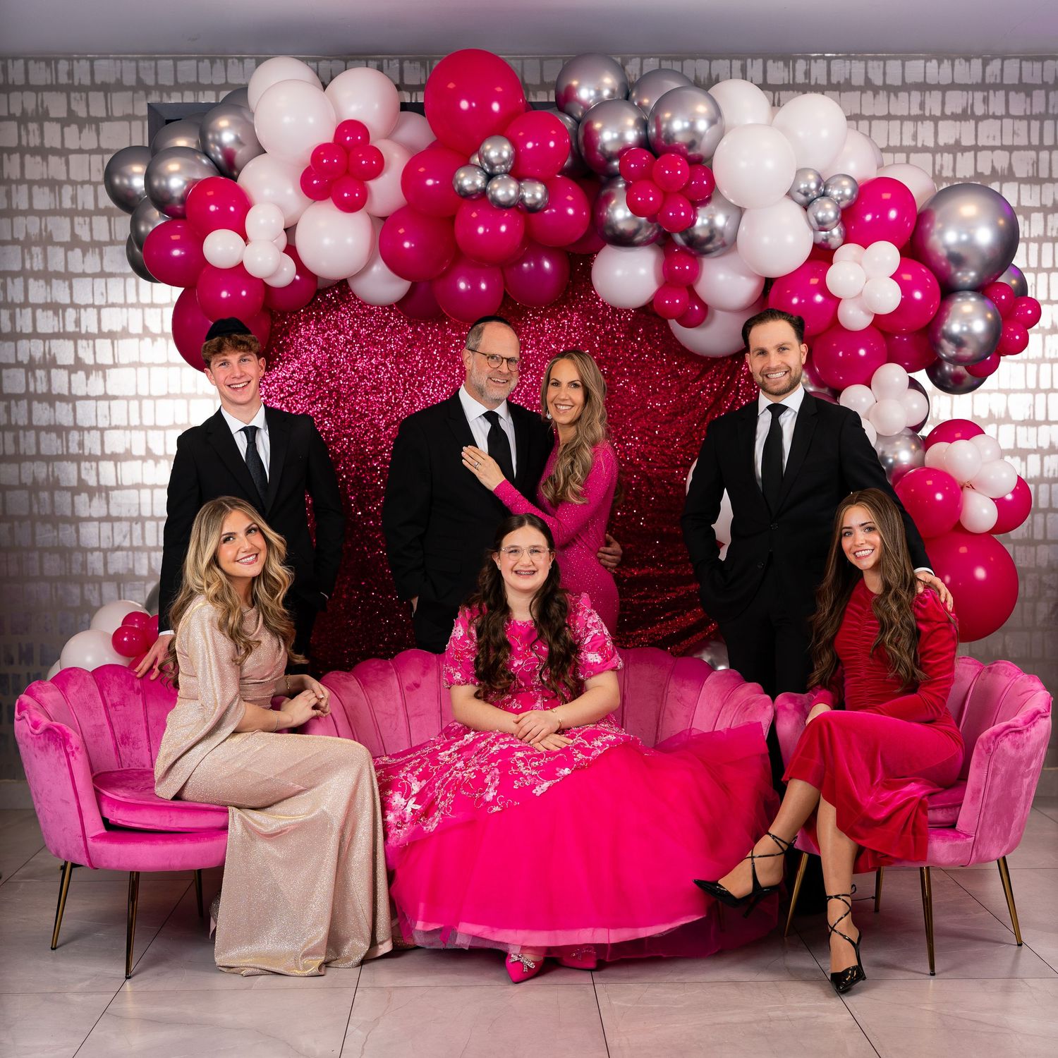 Family group photo in front of pink balloon arch and sequin backdrop, wearing formal attire in pink and black tones.
