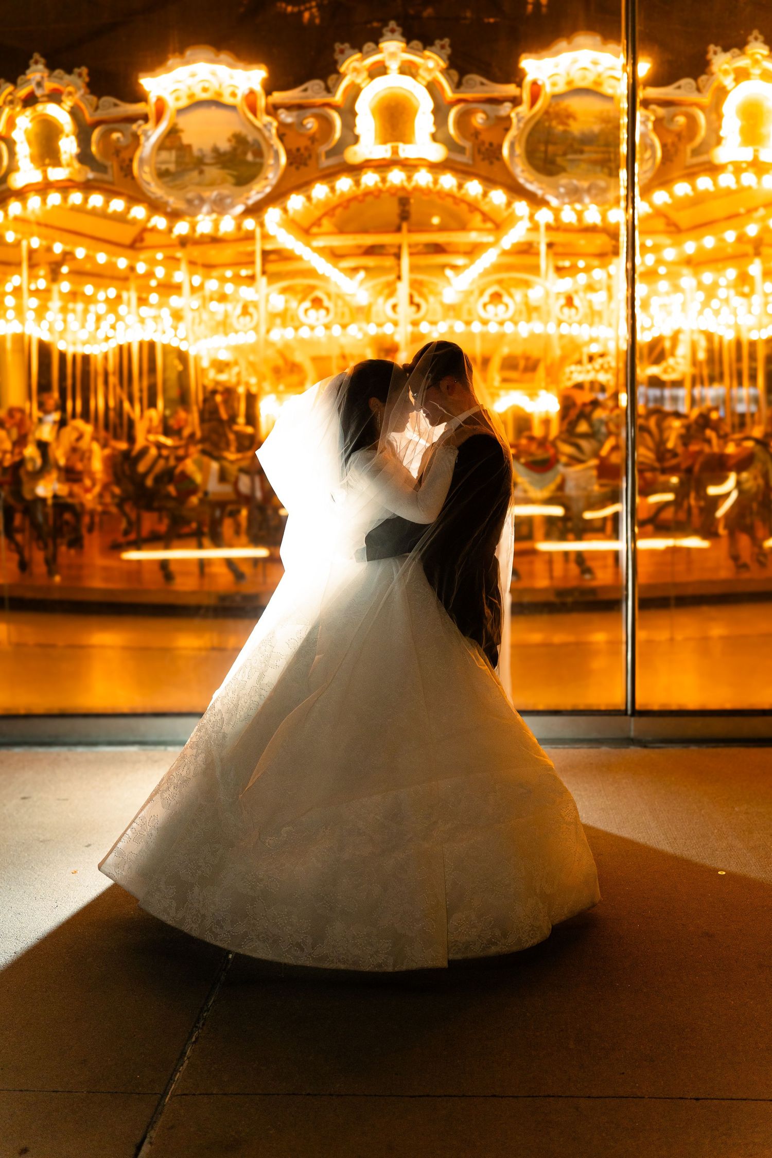 A couple in wedding attire dances in front of an illuminated carousel at night with glowing lights in the background.