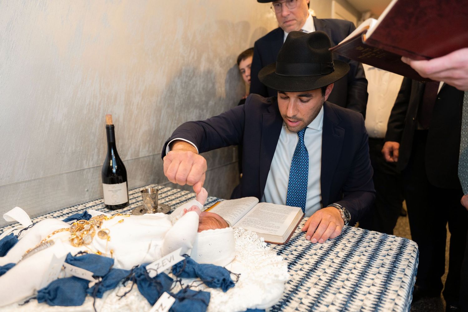 A person in a black hat and blue tie sits at a table with wine and wrapped gifts at a celebration.