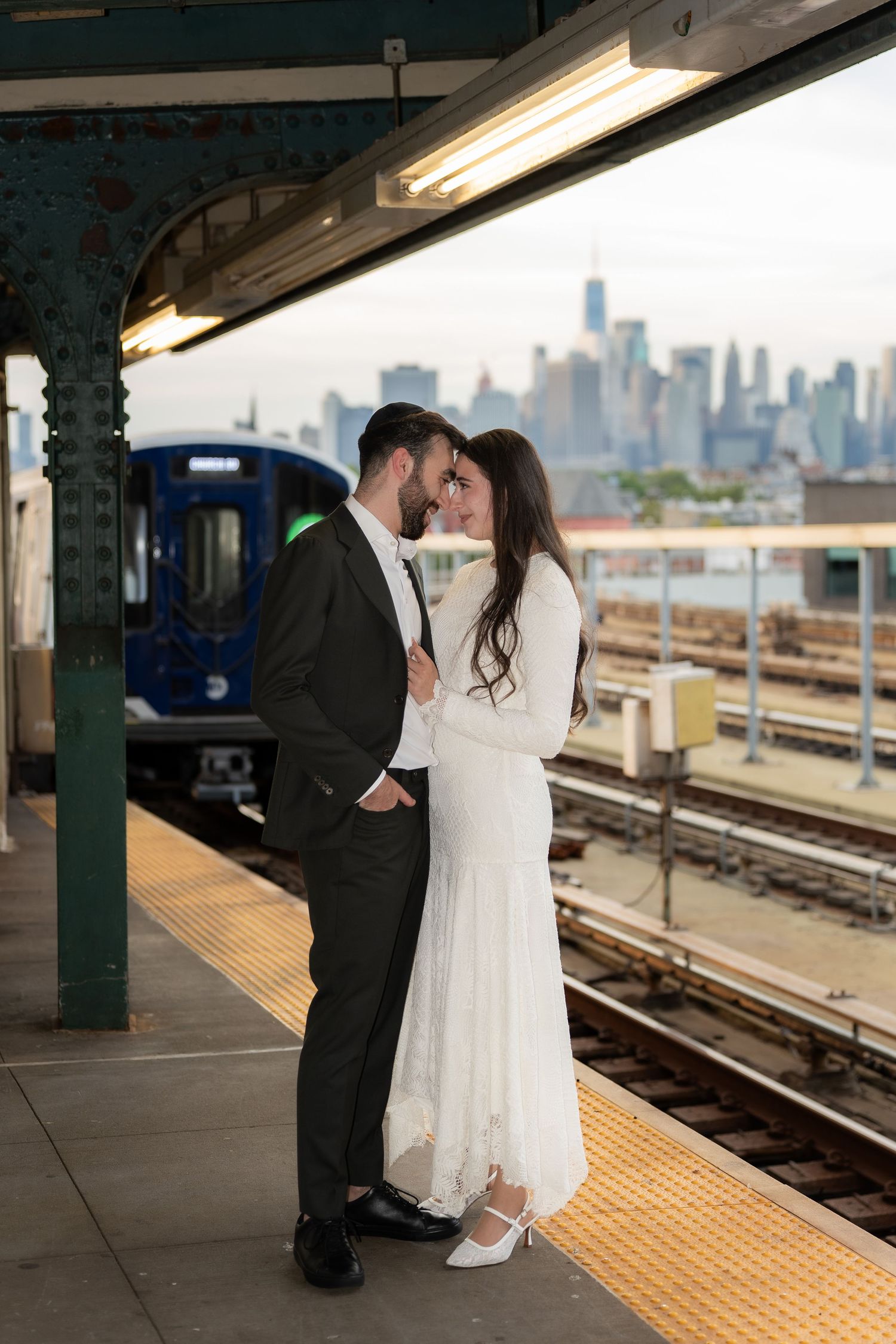 Newlywed couple in wedding attire sharing a romantic moment on a NYC subway platform with Manhattan skyline backdrop.