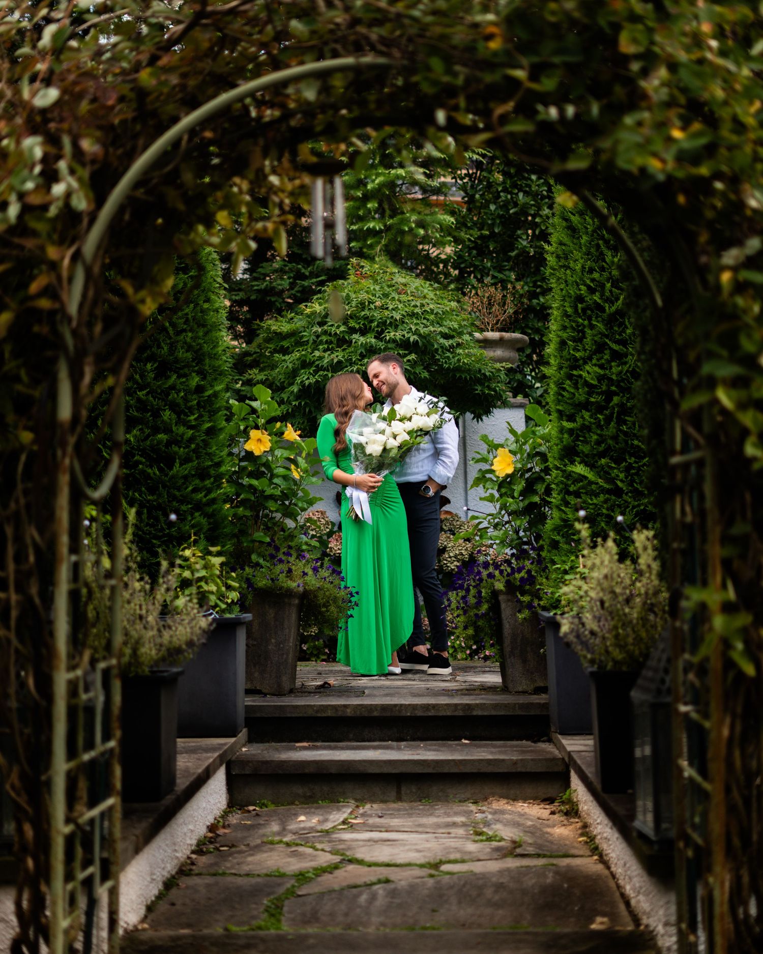 A romantic couple stands beneath a lush garden archway with climbing vines and yellow flowers in bloom.