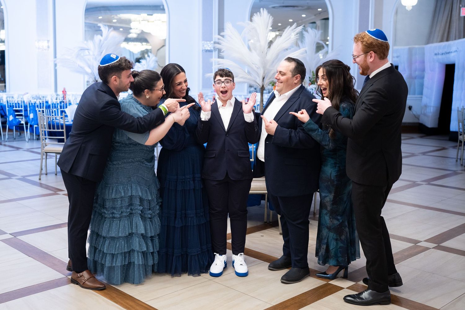 A joyful Bar Mitzvah celebration with family and friends in formal attire gathered around a smiling boy in a decorated ballroom.