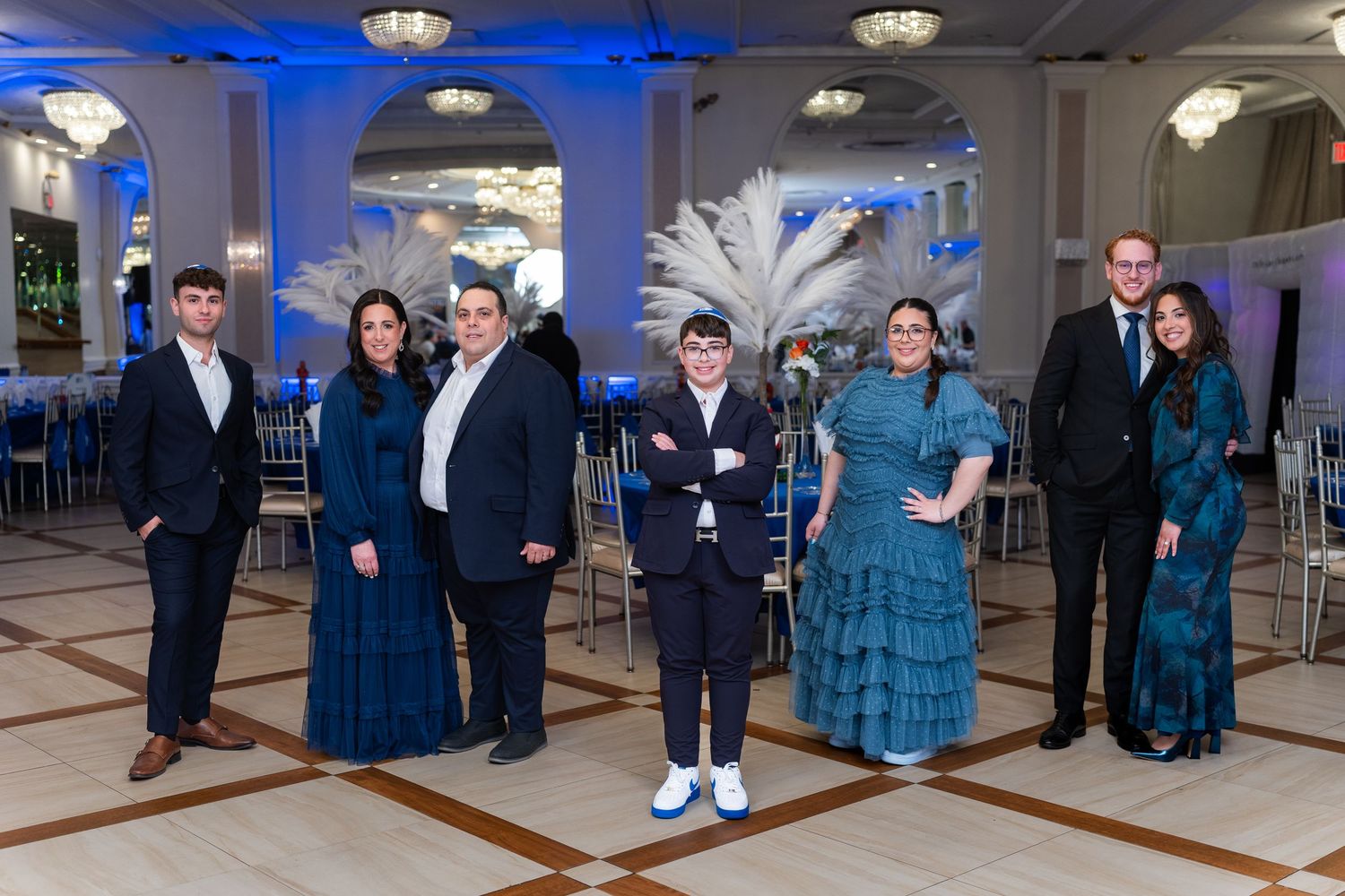 Group of seven people in elegant blue formal attire posing in a decorated ballroom with blue lighting.