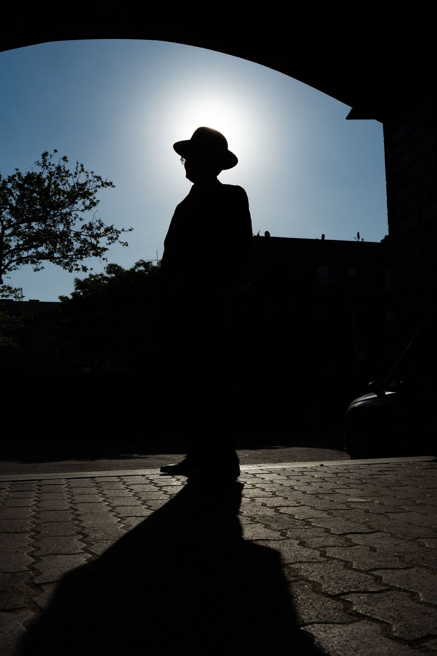 Silhouette of a man wearing a hat standing on cobblestone pavement under an arch with sun glowing behind him.