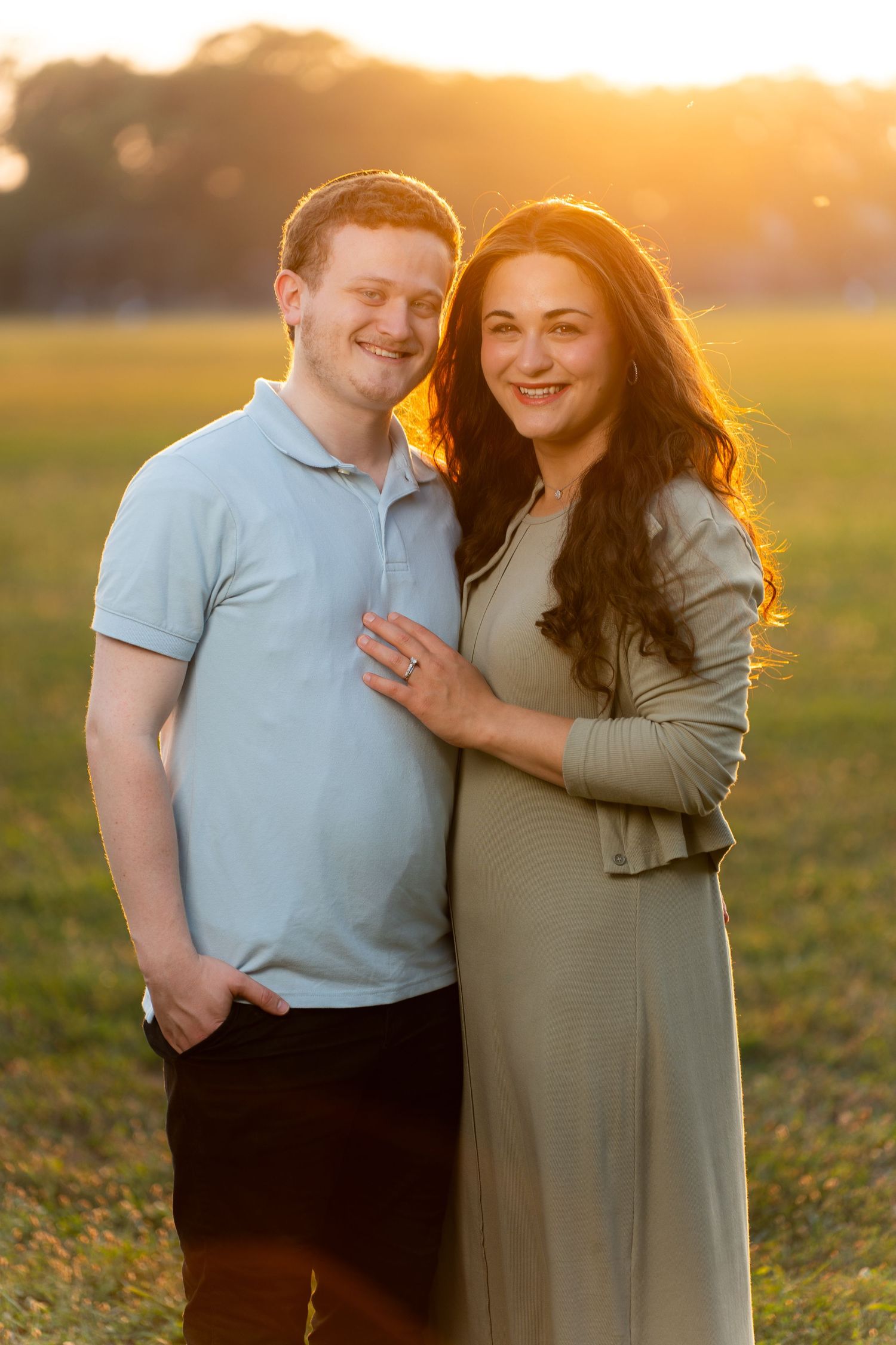 A couple poses together in a field during golden hour sunset with warm backlighting creating a romantic atmosphere.