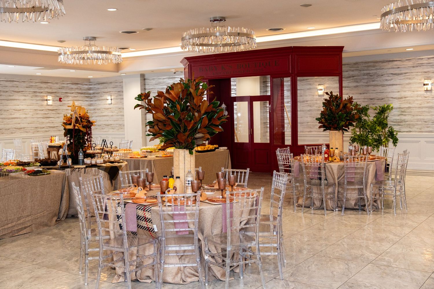 An elegant banquet hall with crystal chandeliers, silver chiavari chairs, and tall floral centerpieces in burgundy and orange.