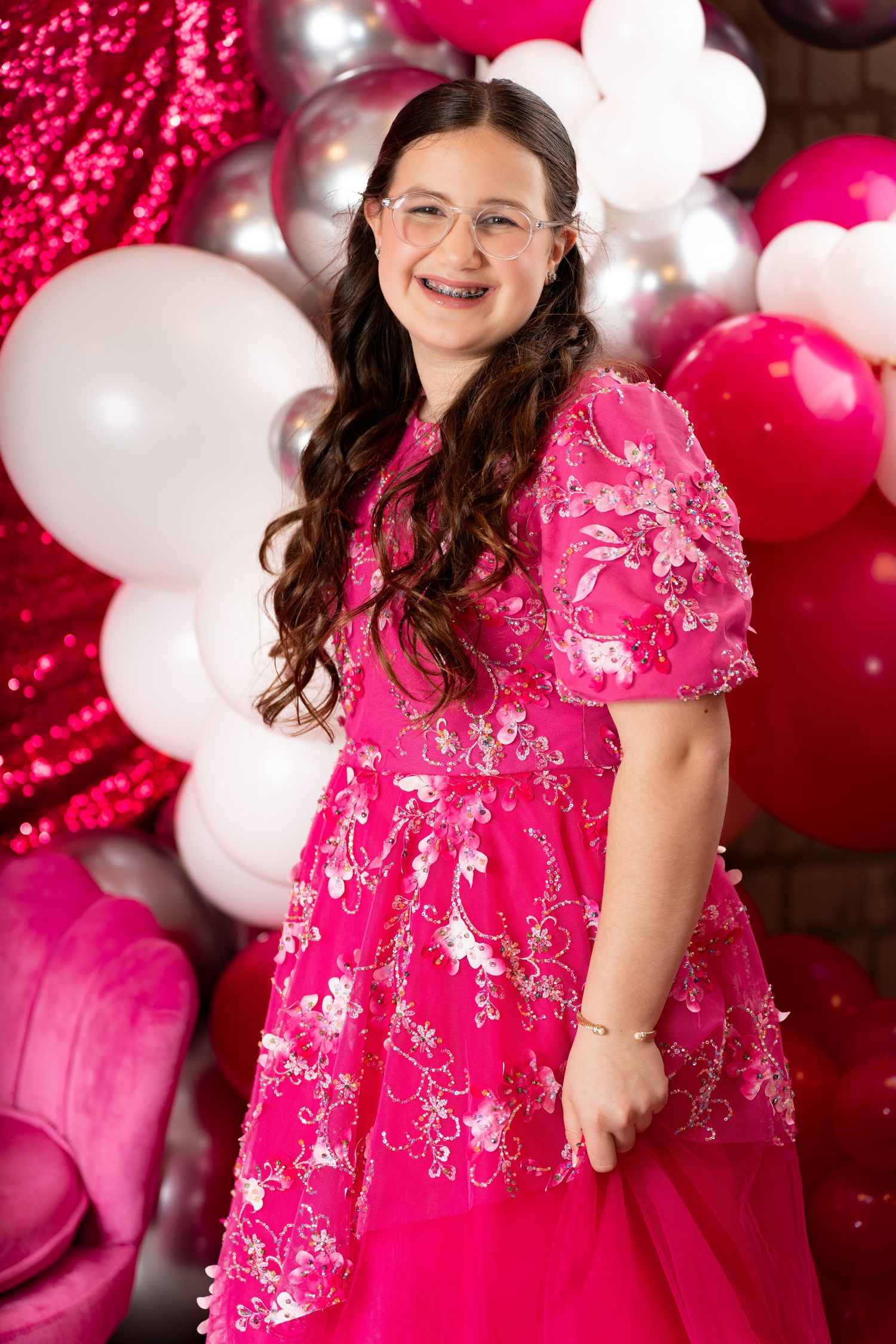 Young girl in pink floral dress with glasses smiles at party with pink and white balloon backdrop.