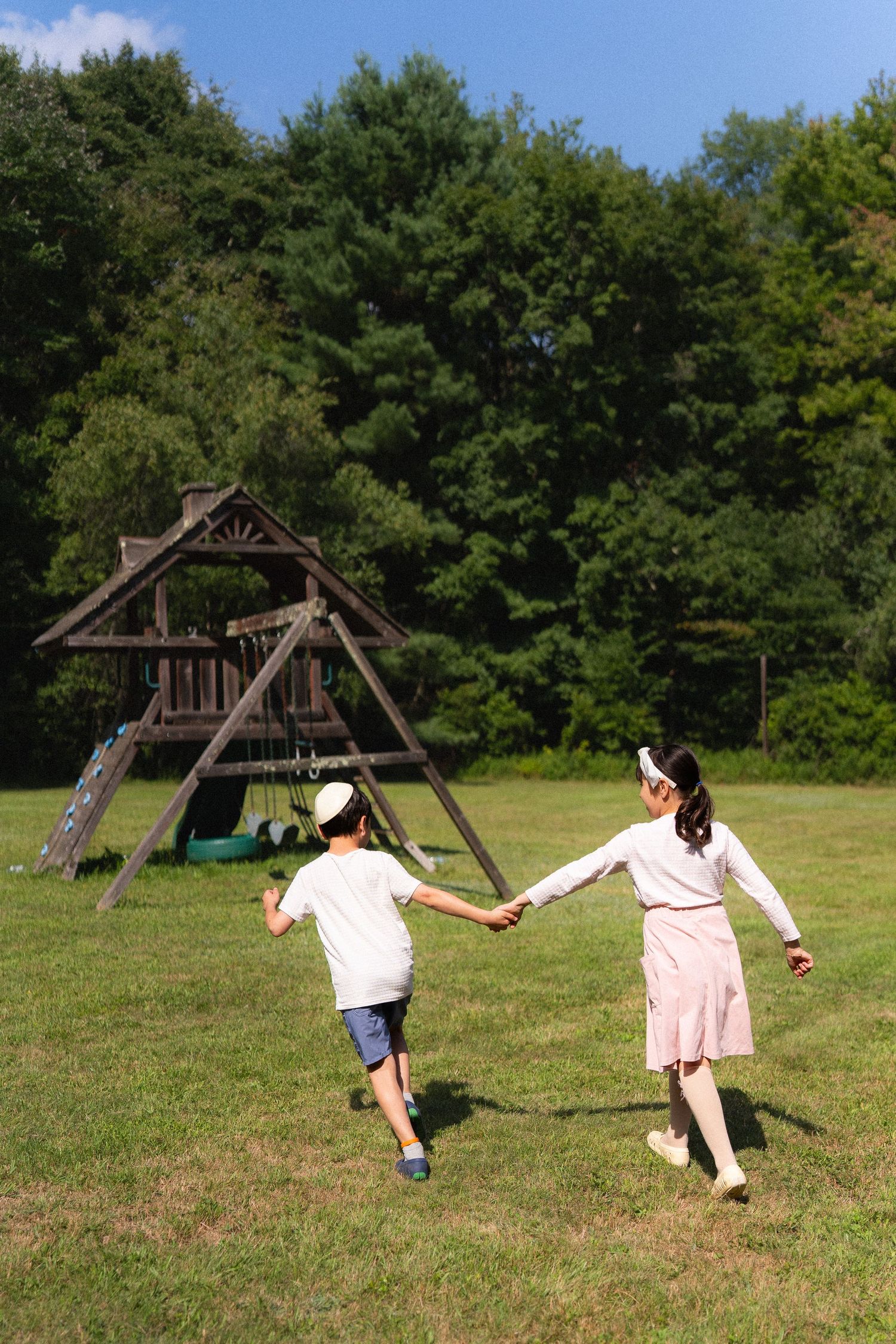 Two children holding hands run toward a wooden playground set on a sunny green lawn surrounded by trees.