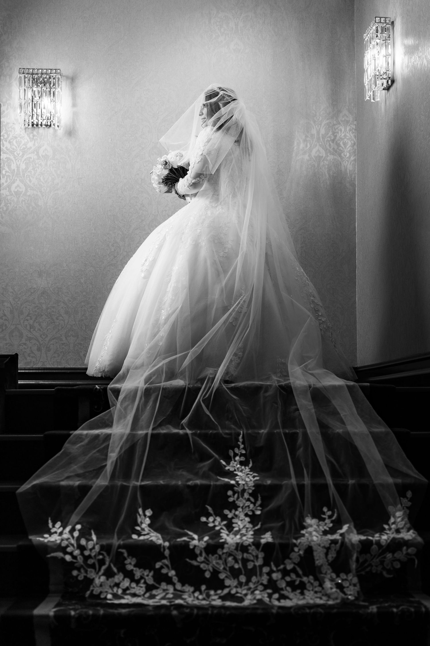 A dramatic black and white portrait of a bride in a flowing veil standing on elegant stairs.