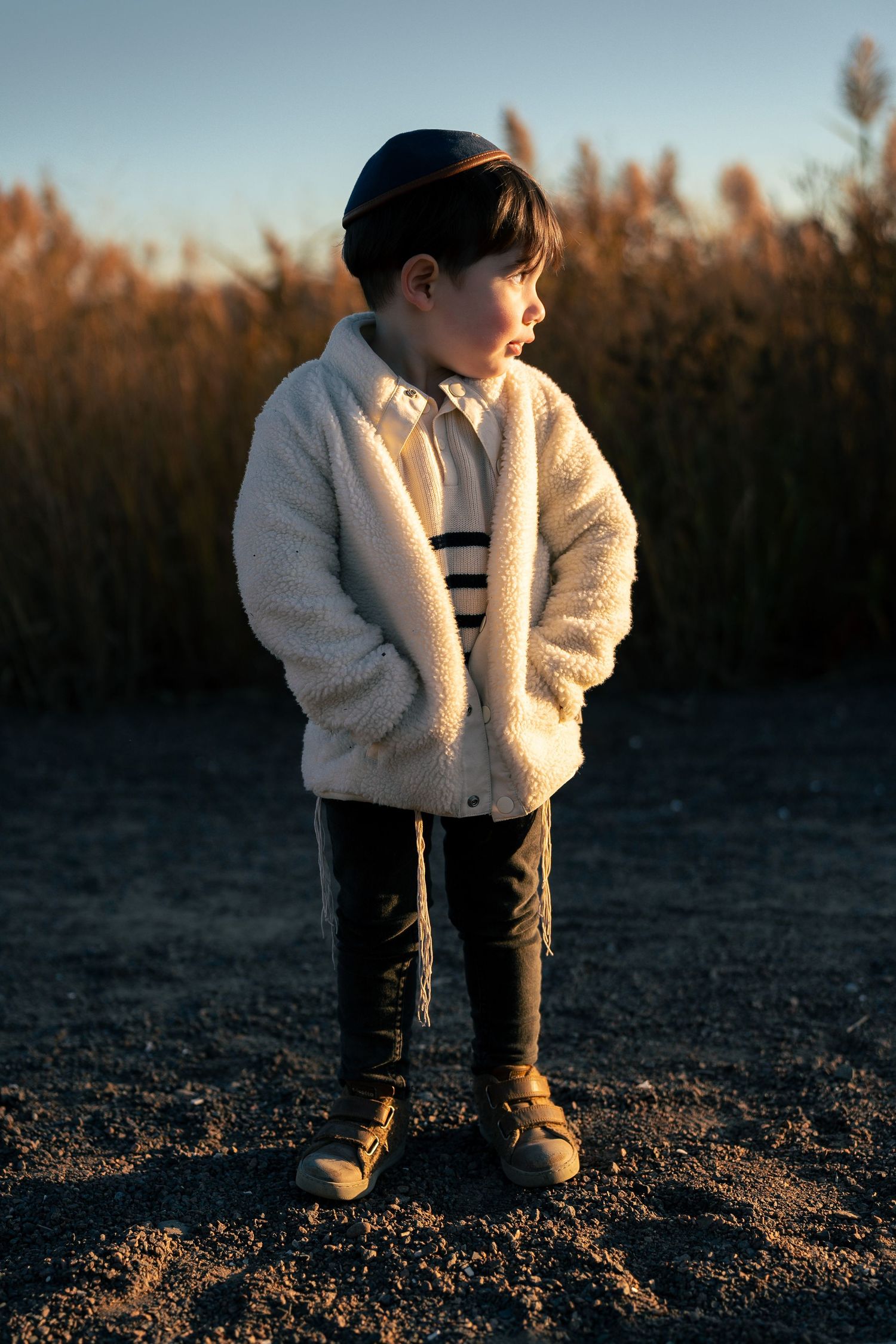 A child in a white fleece jacket and dark pants stands against a sunset backdrop of tall grass.