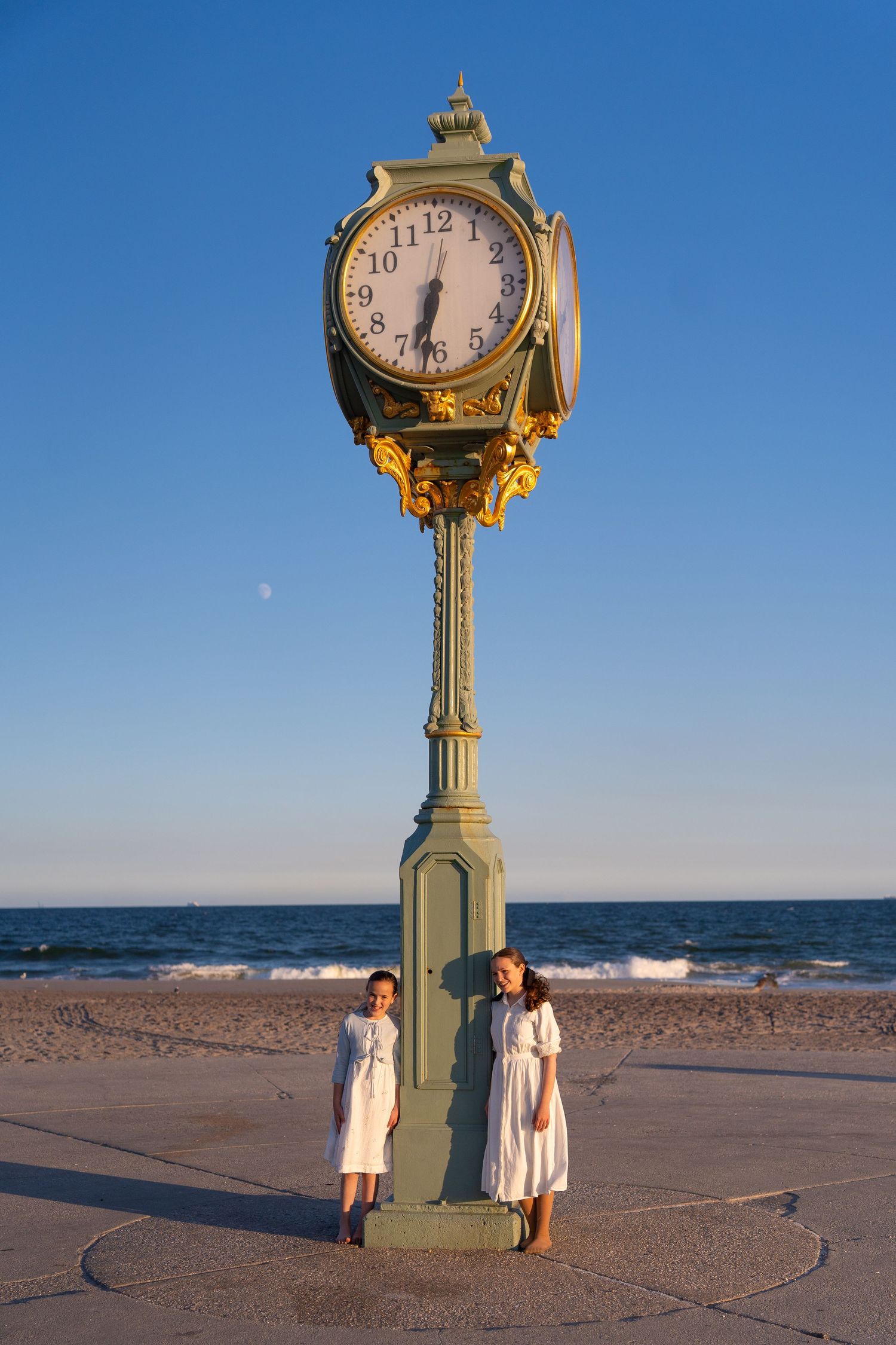 Vintage clock monument stands on beach at sunset as people gather around to admire the seaside timepiece.